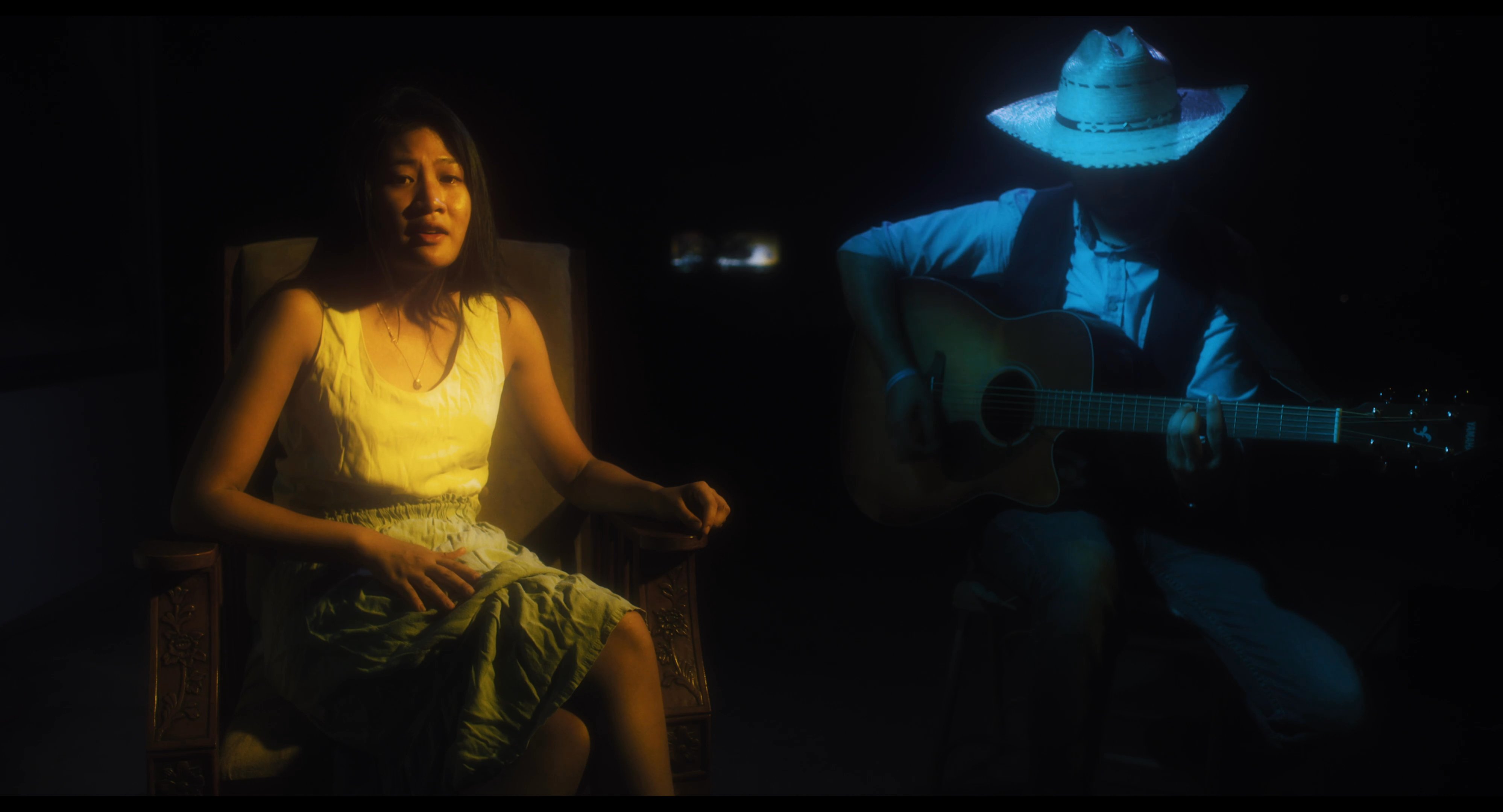 A female vocalist performs in a dimly lit studio while accompanied by a guitarist wearing a cowboy hat. The duo delivers an emotive musical piece highlighted by close-up shots of the singer's expressive performance.