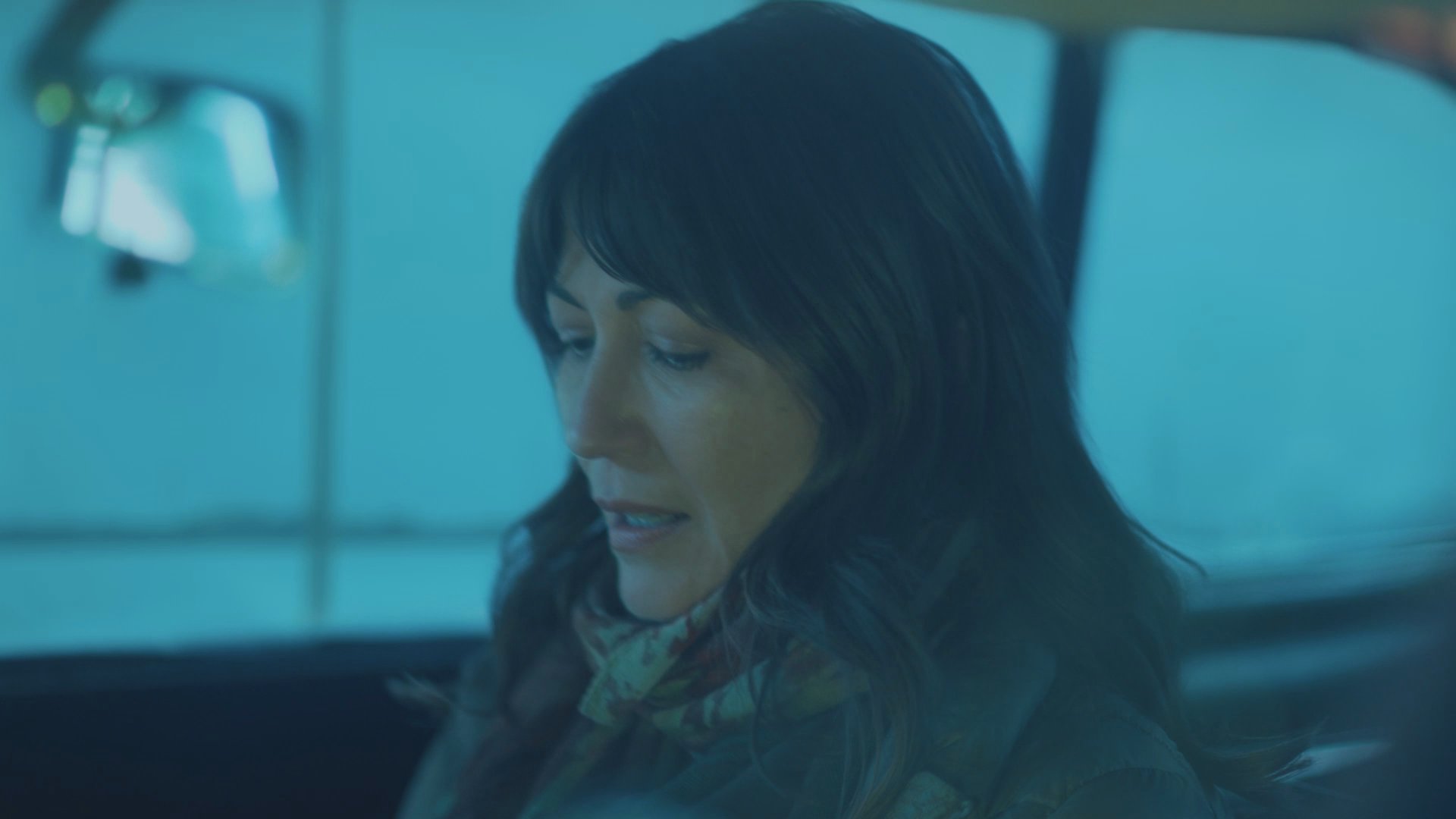 Two individuals engage in a quiet conversation while seated inside a moving vehicle. One passenger checks a smartphone as the pair travels through a landscape illuminated in cool, blue tones.