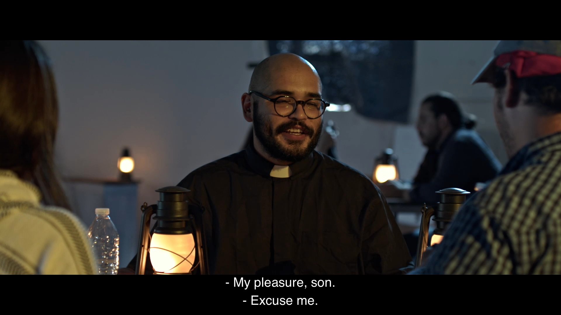 A priest provides hospitality to travelers in a dimly lit room illuminated by lanterns. The interaction culminates in a formal blessing as the group prepares to depart.