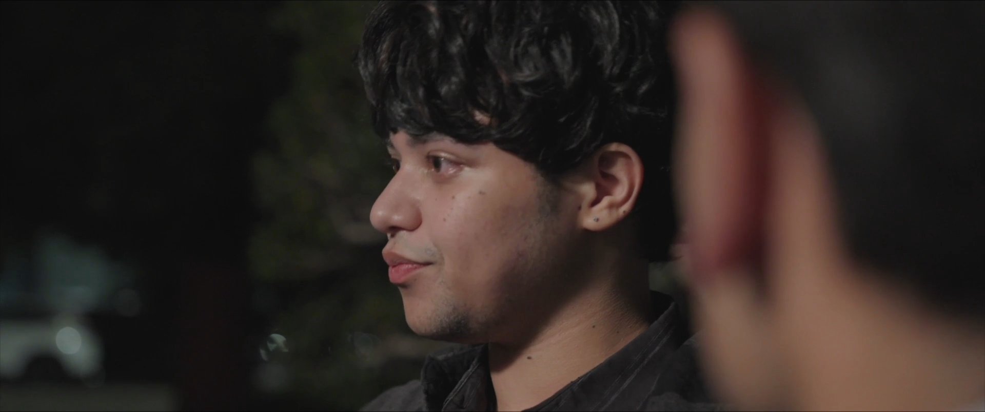 Two men engage in a serious, quiet conversation while illuminated by soft lighting against a dark evening background.