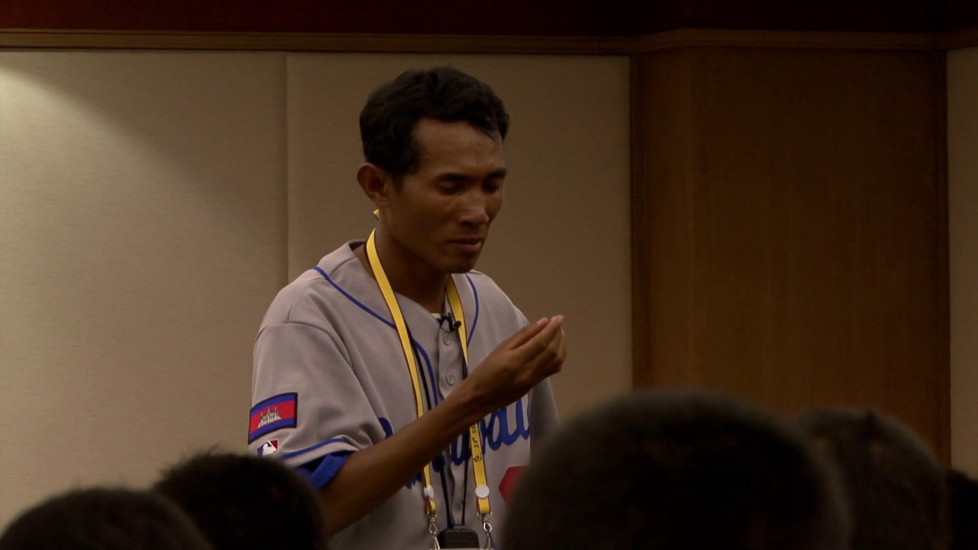 A frustrated baseball player in a Cambodia team uniform voices his grievances to a group during an indoor meeting, questioning the team's performance and reliability.