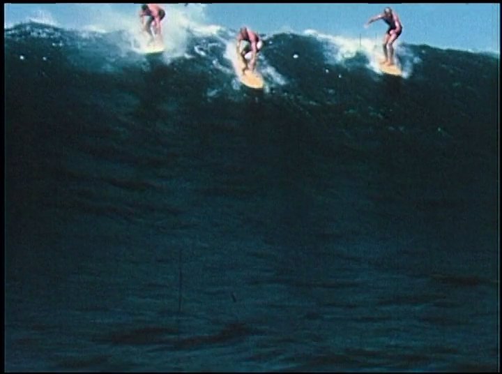 An experienced surfer recounts his memories while archival footage displays men riding large ocean waves on vintage surfboards.