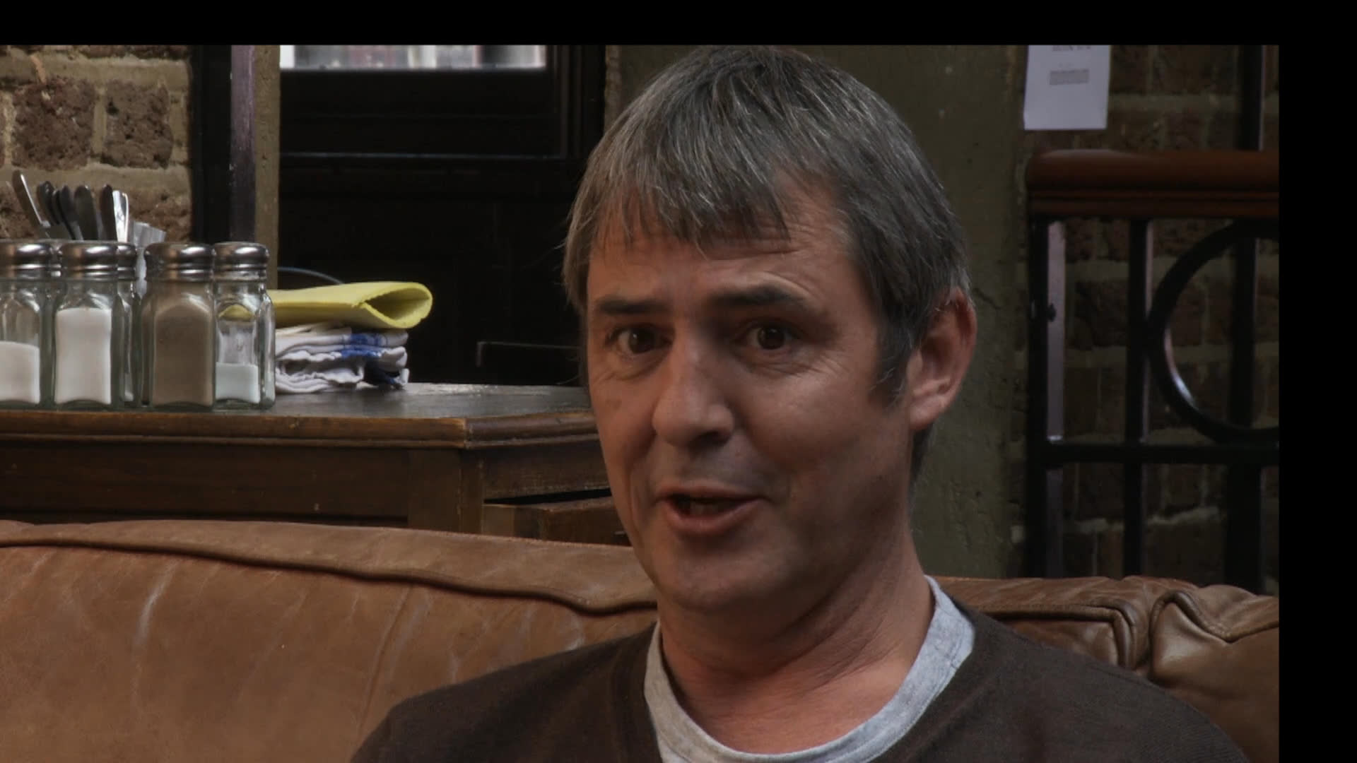 Two men sit in a pub setting, engaged in a conversation while one reads a newspaper. They alternate between speaking and listening, maintaining a relaxed atmosphere throughout the exchange.