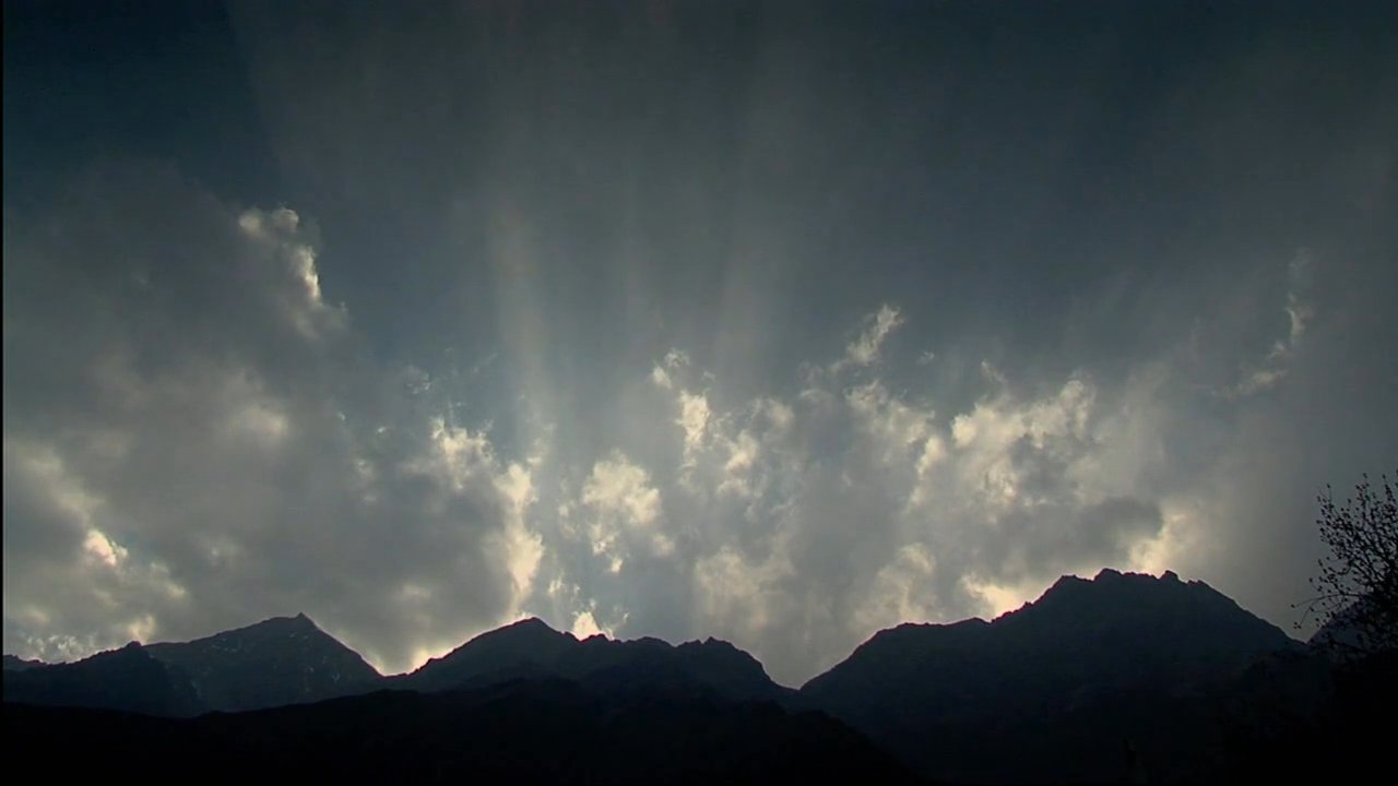 Sunlight breaks through clouds over a silhouetted mountain range, creating dramatic crepuscular rays before shifting to a clear view of snow-capped peaks adorned with prayer flags.