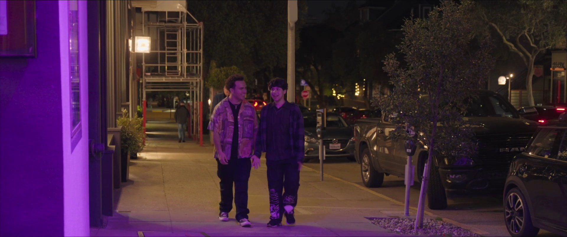 Two young men engage in conversation while standing in a doorway, before transitioning to a walk together along a city street at night illuminated by purple ambient lighting.