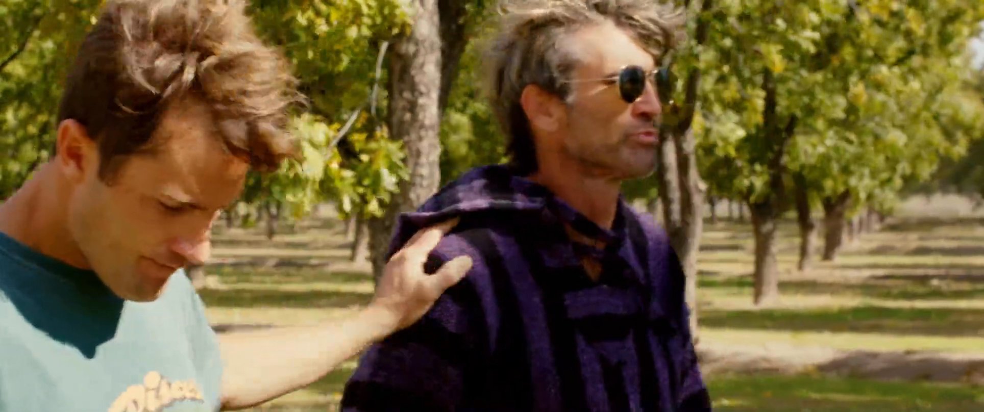 A younger man engages in a supportive walk through a sunny orchard with an older individual wearing sunglasses. The pair continues their conversation while moving from the trees toward a patio area.