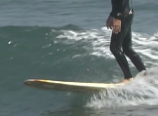 A skilled surfer in a wetsuit maneuvers a longboard across the face of a rolling ocean wave. The athlete maintains balance and momentum while navigating the breaking water.
