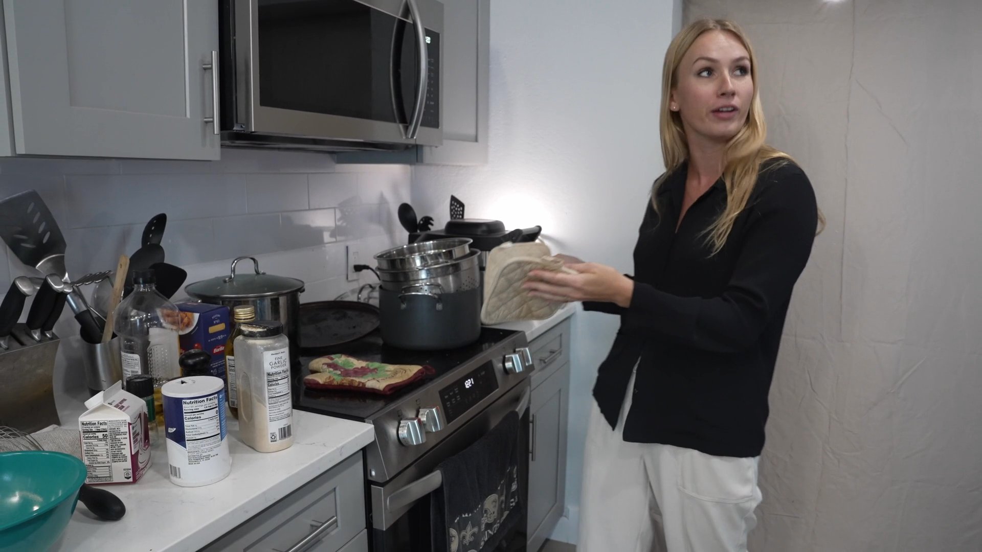 A woman demonstrates cooking techniques in a kitchen setting while two men observe and converse nearby.
