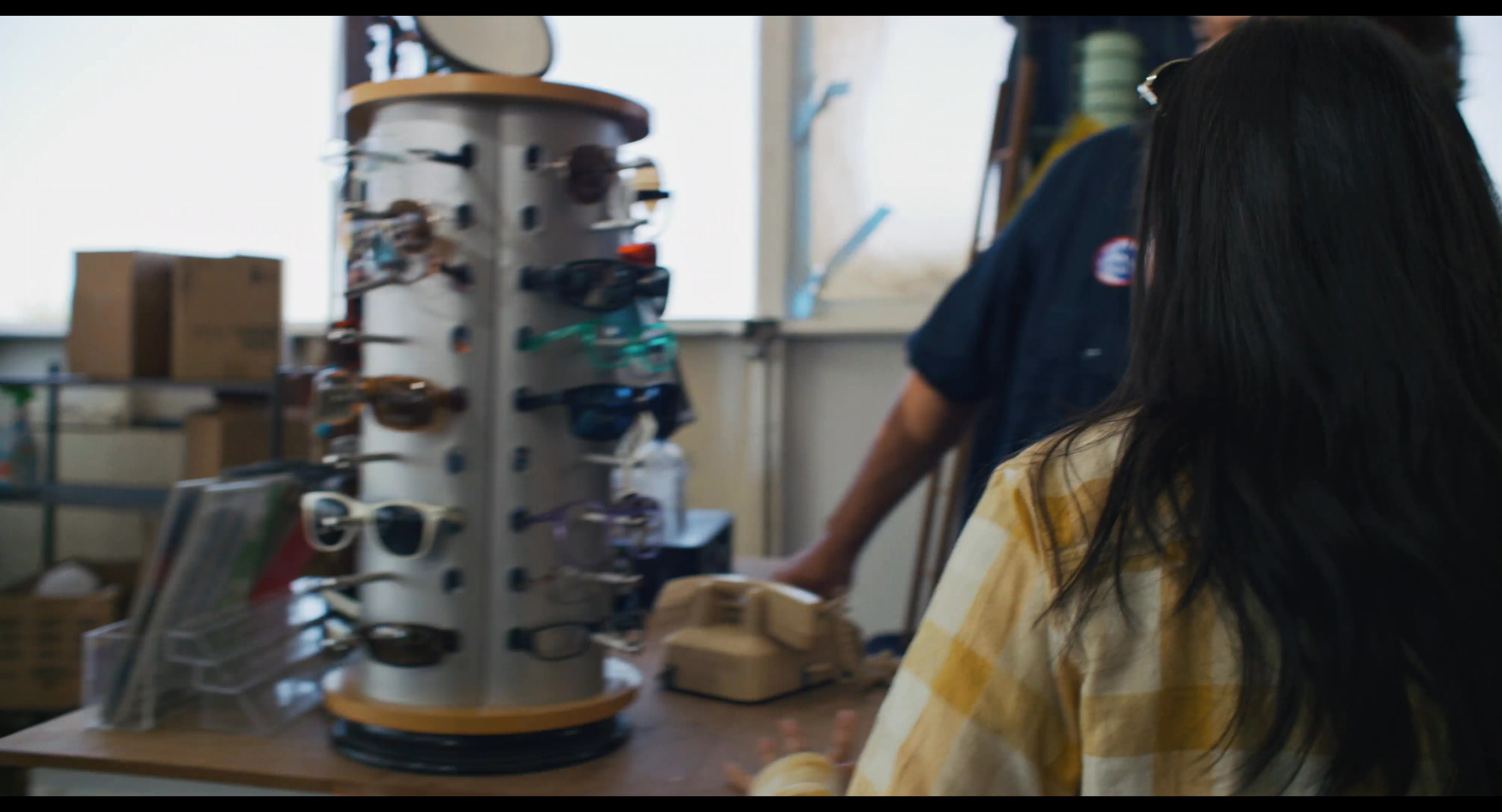 A young traveler carrying a blue suitcase enters a remote roadside store and attempts to use a landline telephone while a clerk observes from behind the counter.