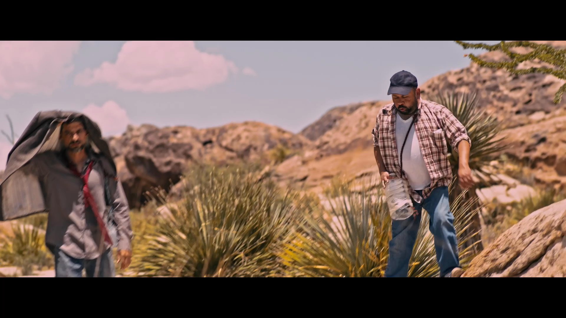Two men navigate a harsh, arid desert landscape while using a jacket for shade against the sun. They continue their journey through the rugged terrain under clear skies.