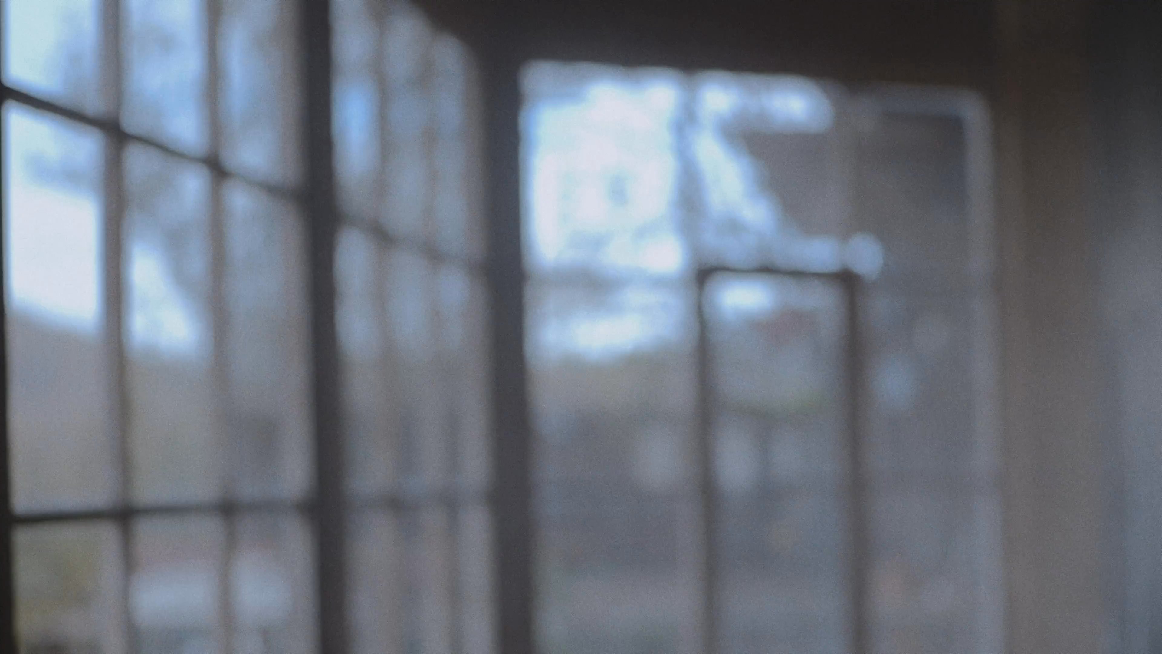 A woman stands in a contemplative state before a man pours water over her head in a deliberate, ritualistic act. The scene takes place against a minimalist architectural backdrop, conveying a somber and transformative atmosphere.
