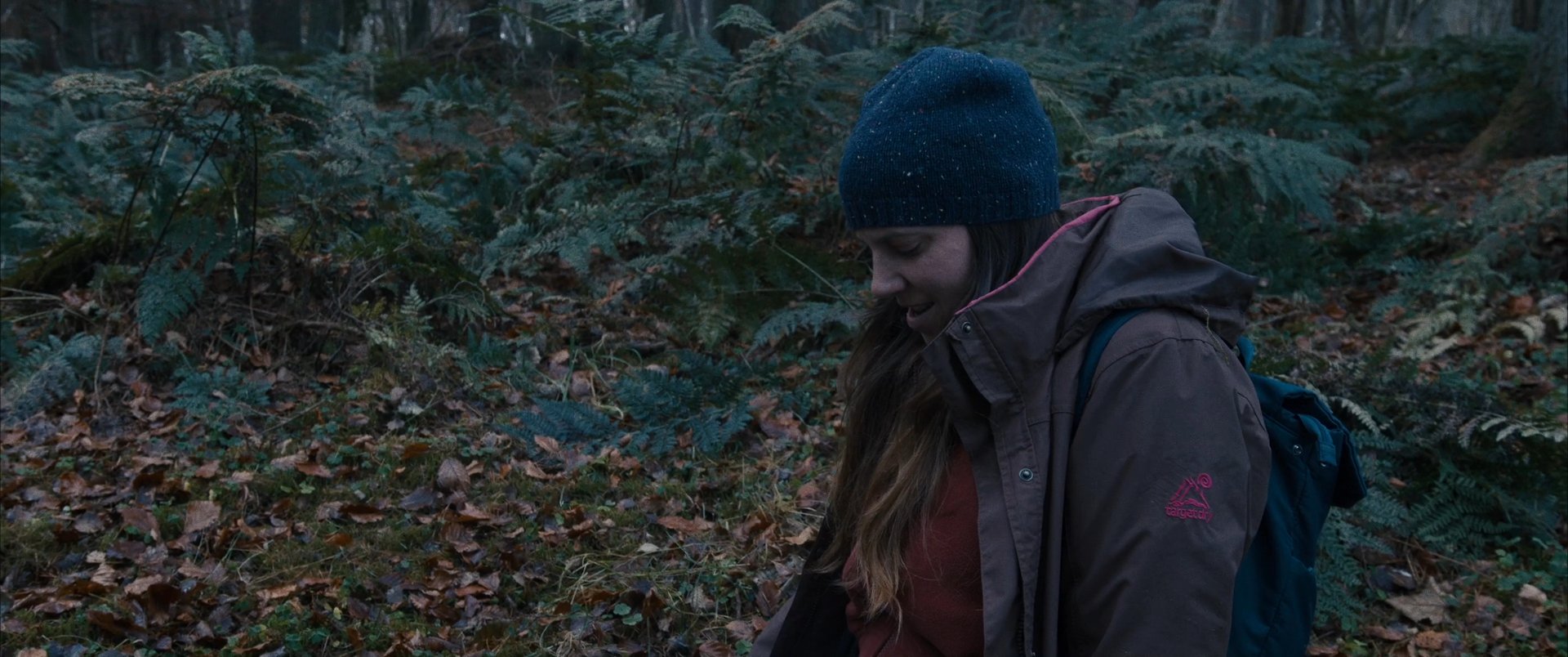 A woman explores a dense woodland, alternating between carrying a bag and navigating the fern-covered terrain in autumn.