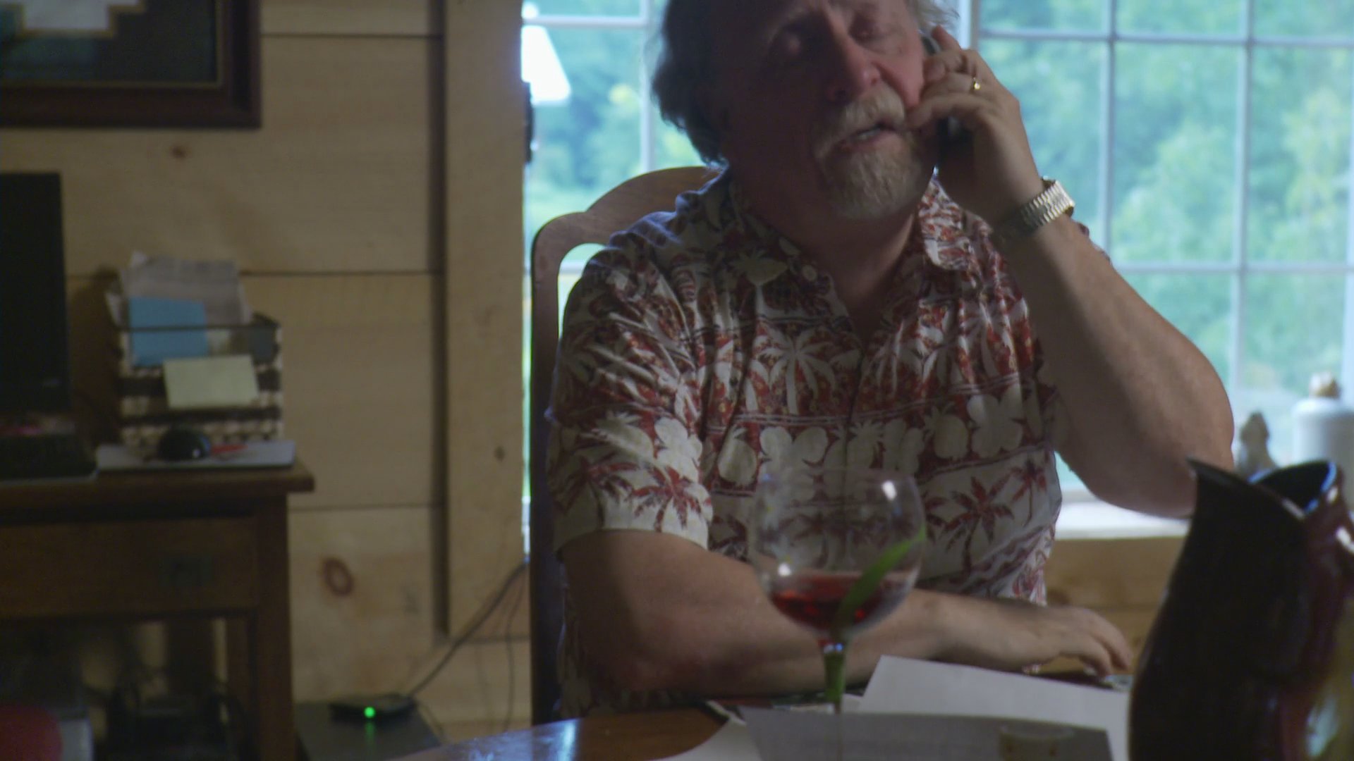A senior man conducts a professional phone call from his home office while a younger woman manages a separate conversation in the background.