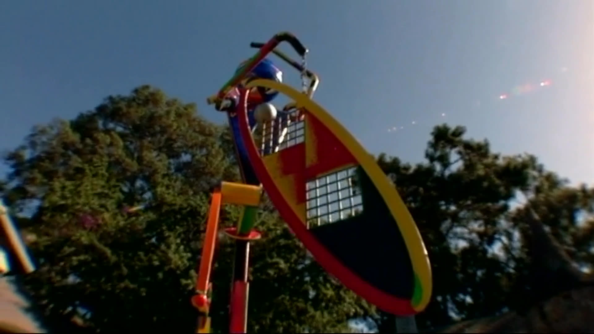 A brightly colored outdoor sculpture with a human-like figure shifts through a series of positions beneath a clear sky. A viewer in a green cap watches as sunlight catches the artwork against a backdrop of trees.
