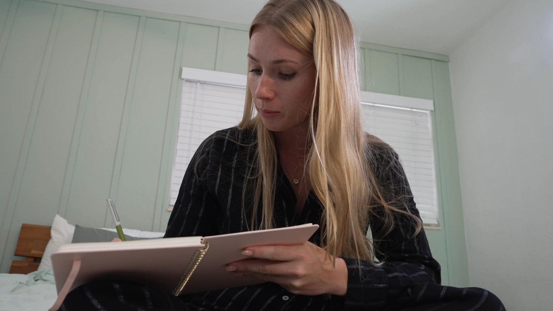 A woman sits in a bedroom and thoughtfully journals, alternating between reflective pauses and writing in her notebook.