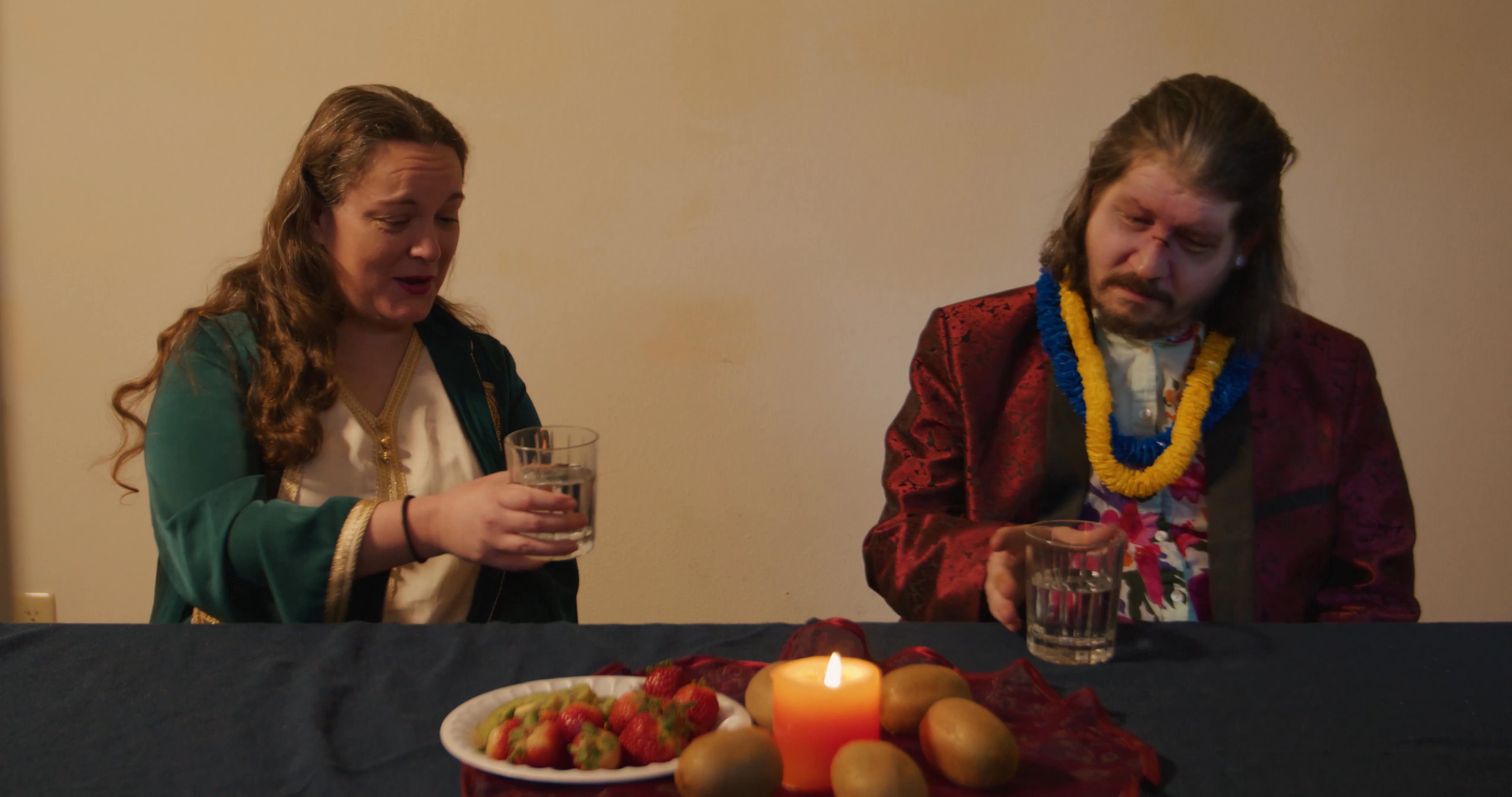 Friends participate in an informal dinner gathering, sharing drinks and conversation at a table illuminated by candlelight. Attendees engage in lighthearted interaction, including the playful distribution of bananas.