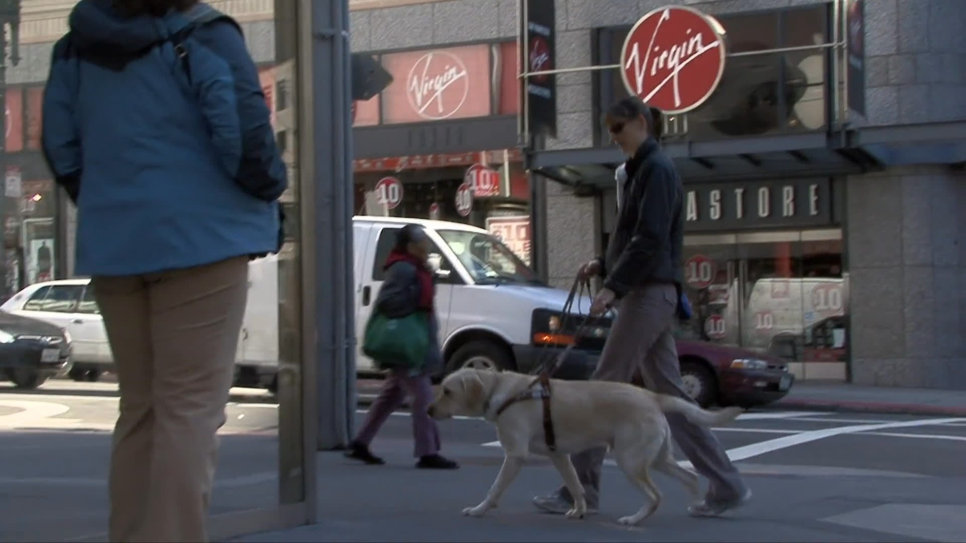 A yellow labrador guide dog assists its owner through a San Francisco BART station entrance and onto an escalator. The pair demonstrates skilled urban navigation in a busy city setting.