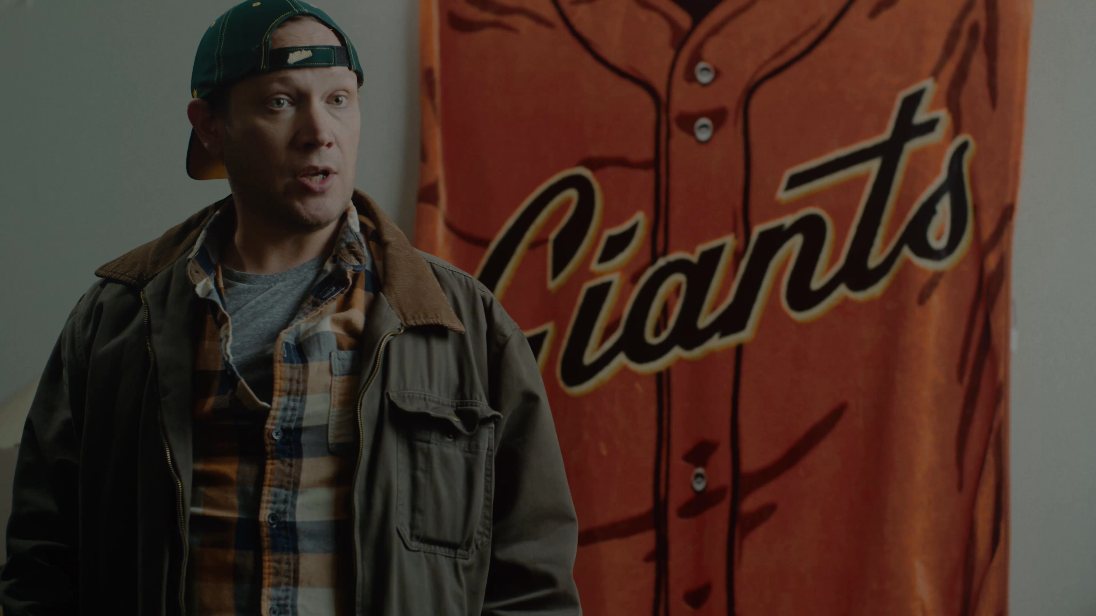 Two individuals engage in a focused conversation, with one subject seated in a yellow armchair and the other standing before a San Francisco Giants blanket, gesturing as they share personal perspectives.