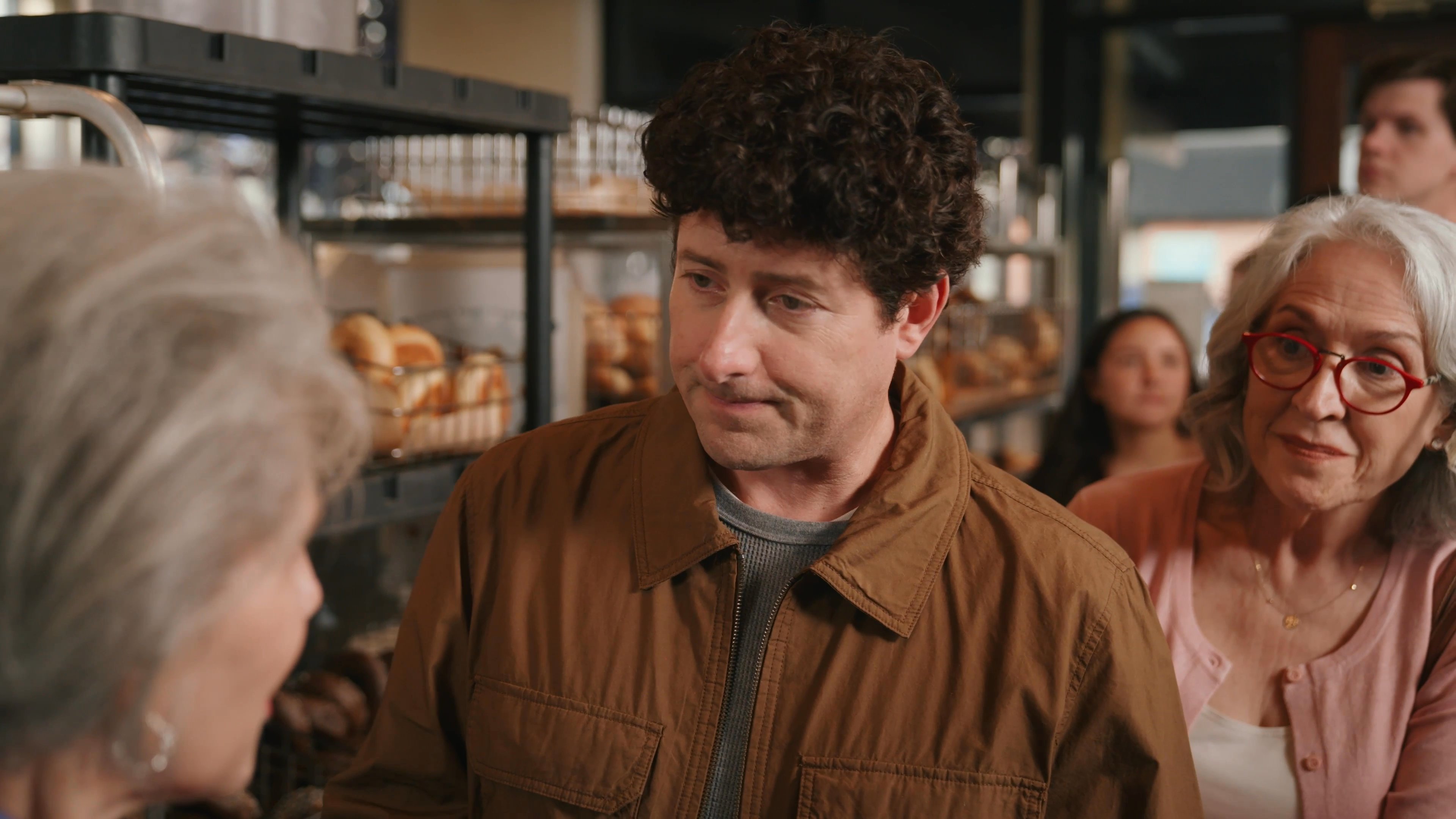 An older woman in business attire engages in a conversation with a man inside a bakery setting. Others appear in the background as the two exchange words in a professional or retail context.