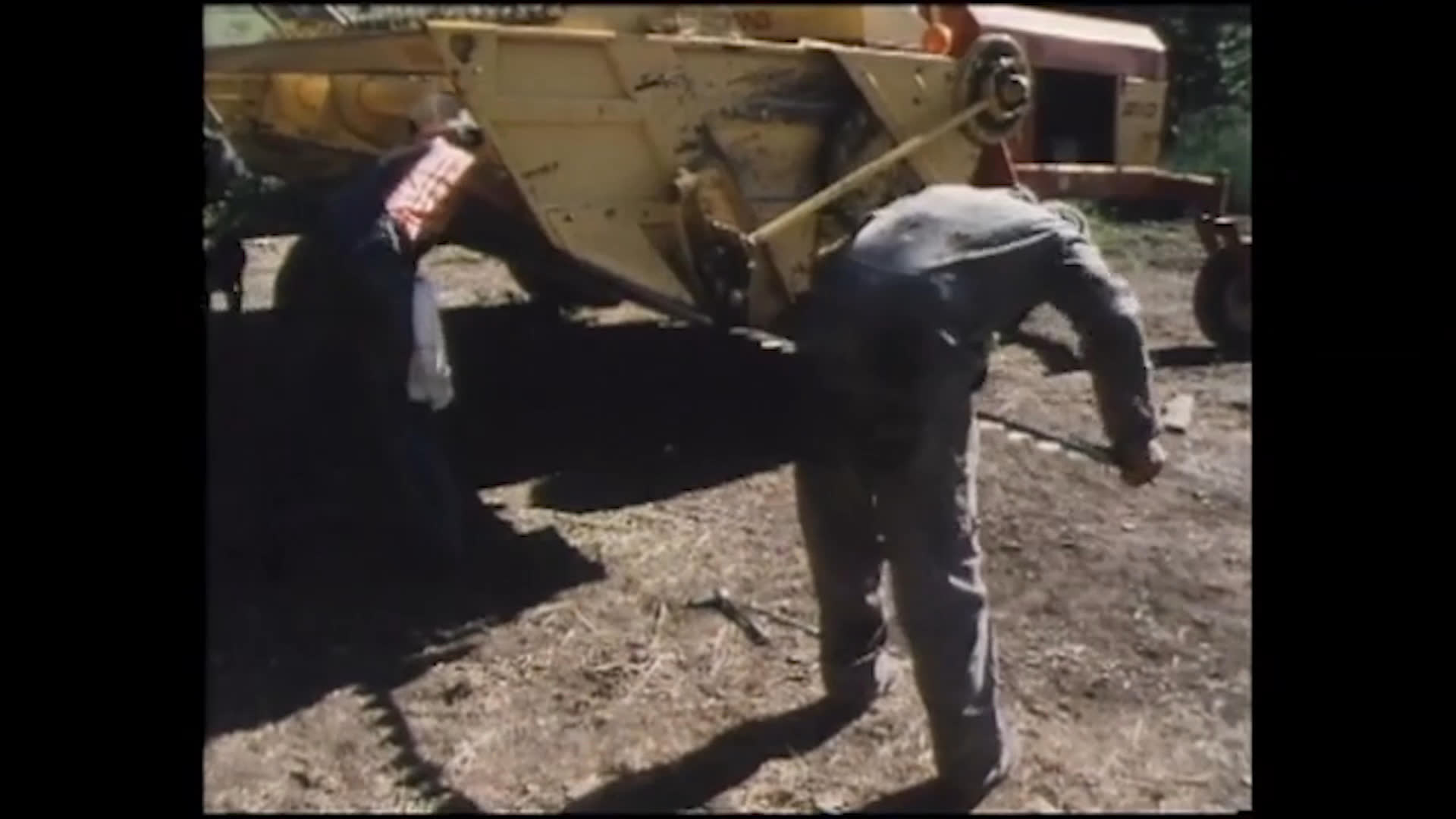 In a sunny farmyard, Bonnie reaches into large yellow agricultural machinery while men work nearby. A man calls her to the job, and another speaks warmly of her as a valued member of the family.
