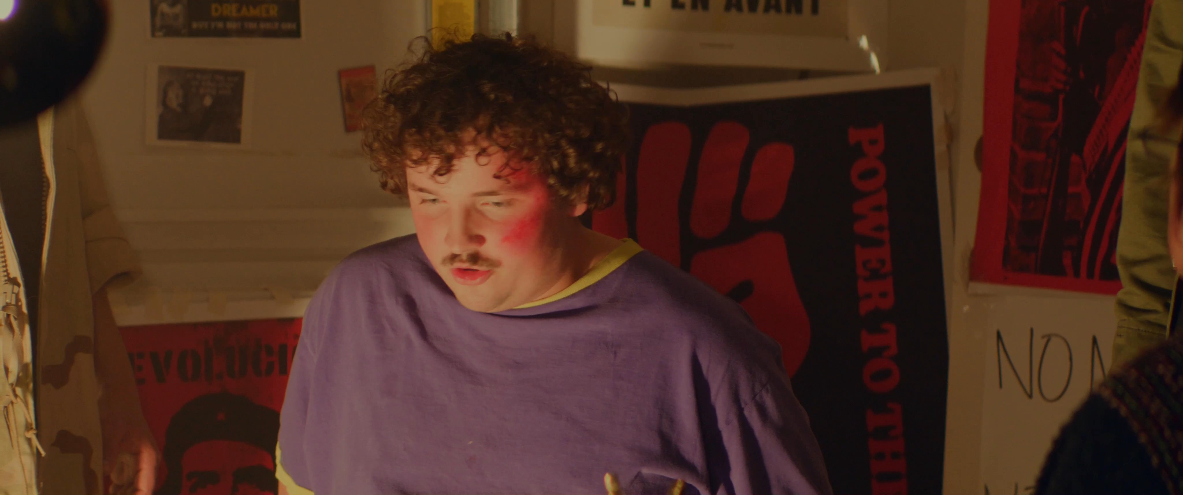 Young individuals engage in a focused, intense conversation within a room decorated with political and revolutionary posters. The atmosphere suggests a clandestine meeting centered on social movements and ideology.
