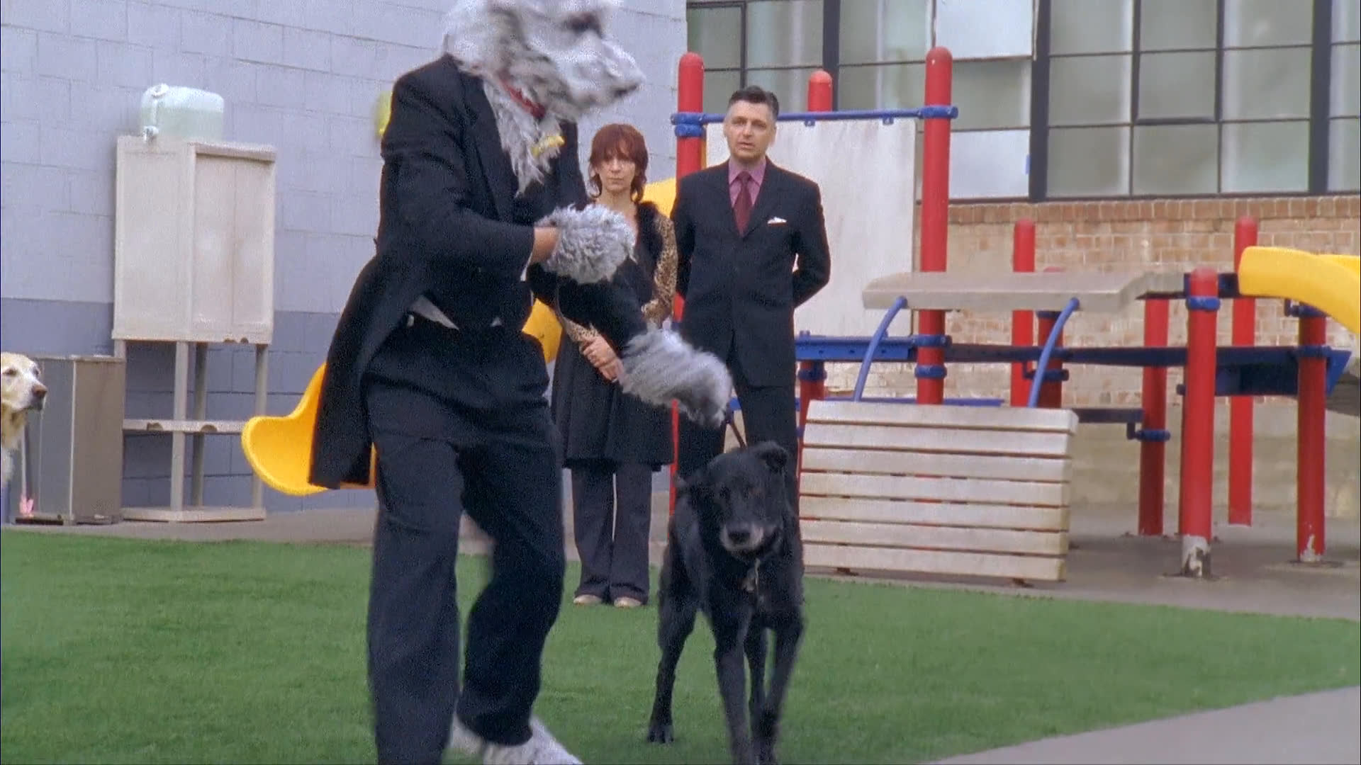 A man wearing a dog costume interacts with a golden retriever in a playground setting before removing the headpiece to reveal himself. The scene highlights a moment of personal transformation in a professional-style attire.