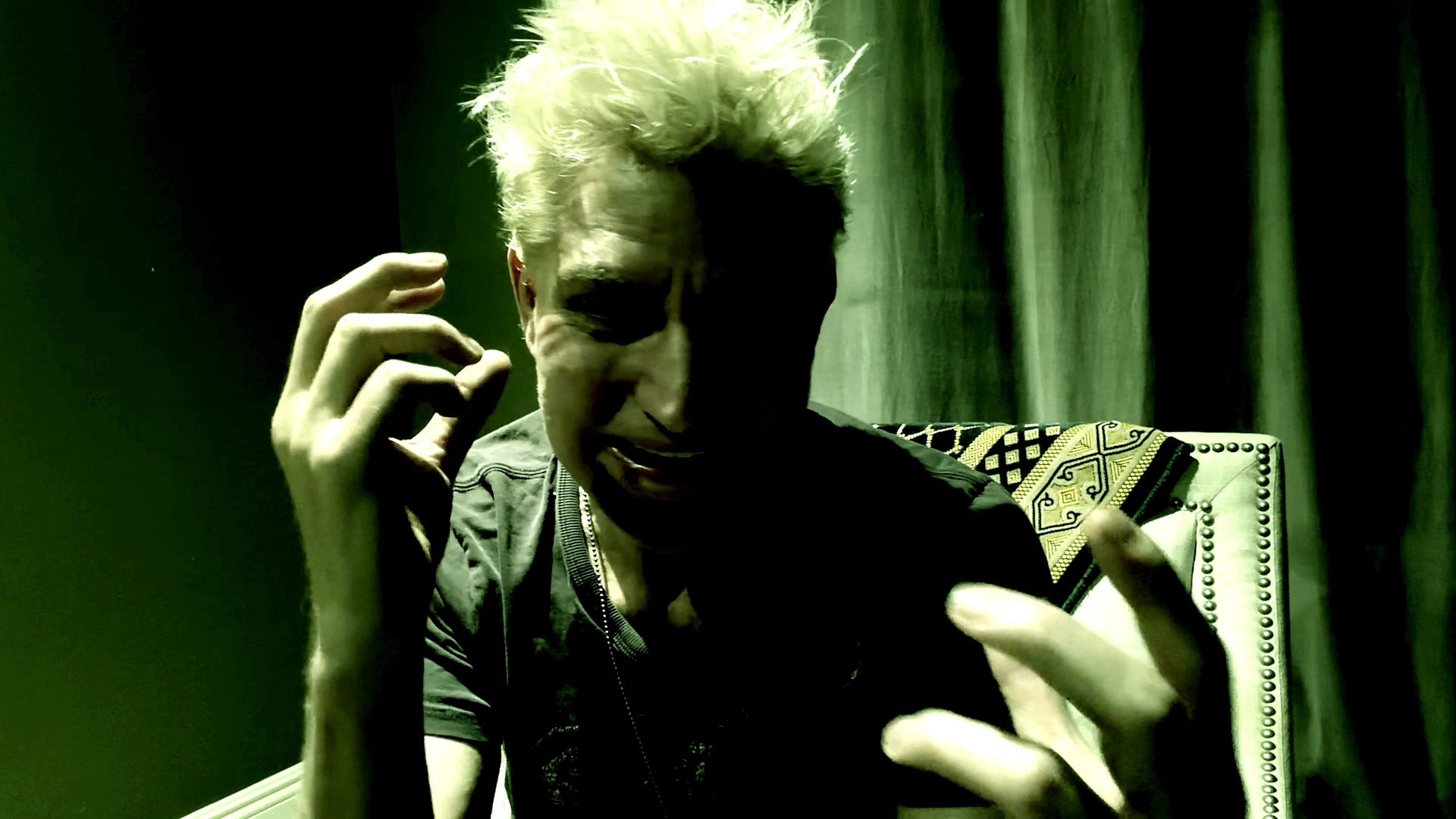 A man exhibits signs of deep emotional turmoil and psychological agitation while speaking directly into the camera. The scene features a stark, green-tinted lighting aesthetic that underscores his intense, dramatic performance.