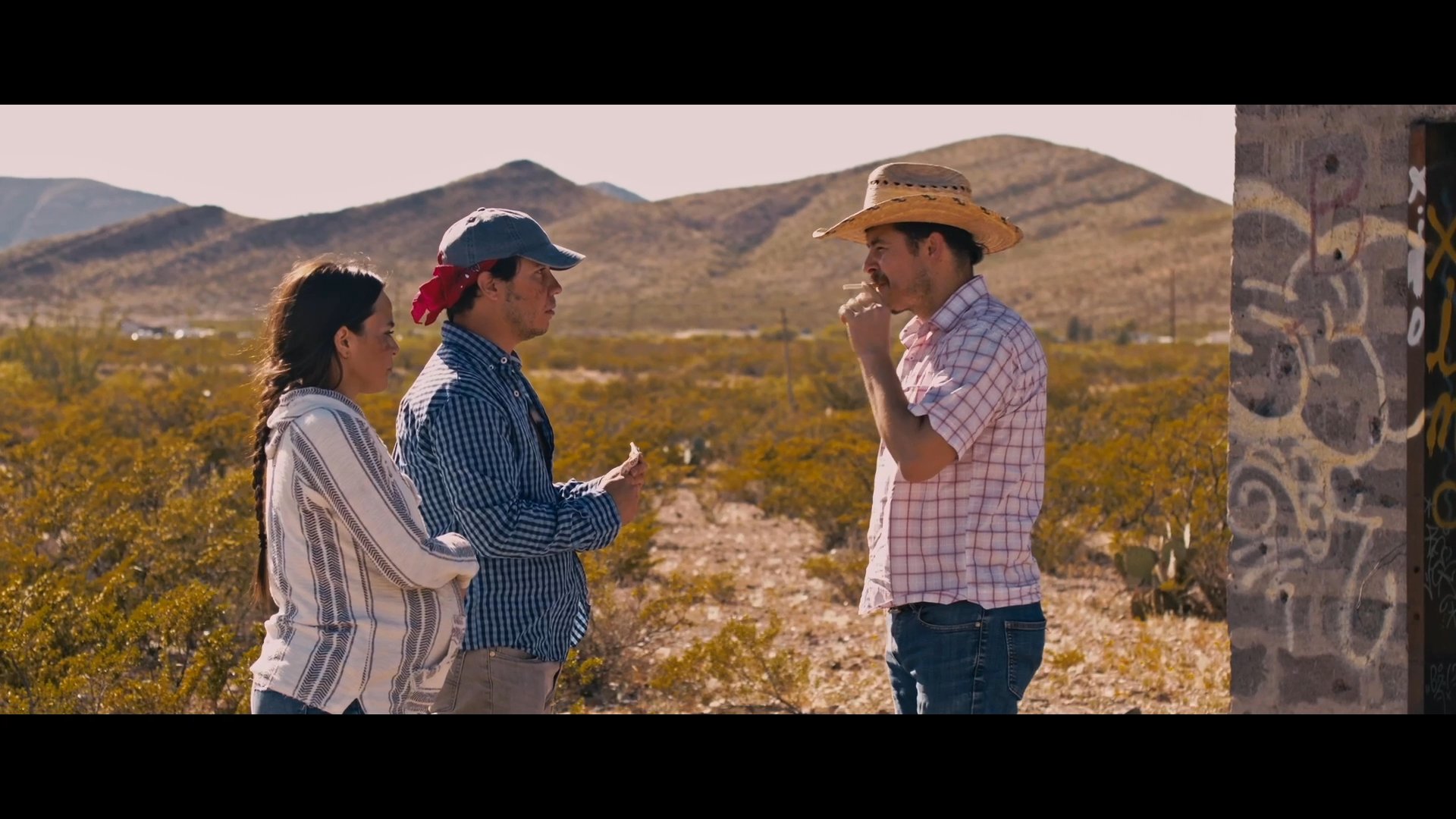 A man in a straw hat questions a couple in the desert about their claim of being robbed. The interaction shifts from a tense financial negotiation to an exchange marked by skepticism and doubt.