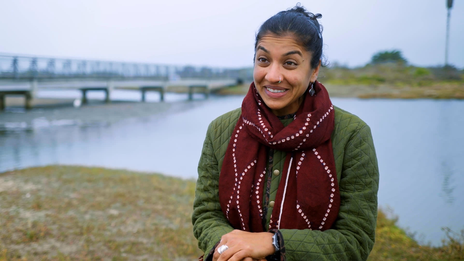 A park representative introduces a newly opened urban national park beside a busy San Francisco highway and waterfront. She discusses community involvement as people prepare a table and lay out park-planning materials on the grass.