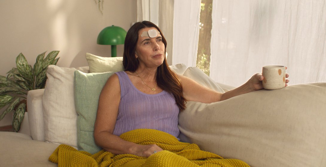 A woman sits on a couch next to a window in a living room. She holds a cup in her hand, has a blanket over her lap, and has the Enso from Hinge Health device on her forehead. 
