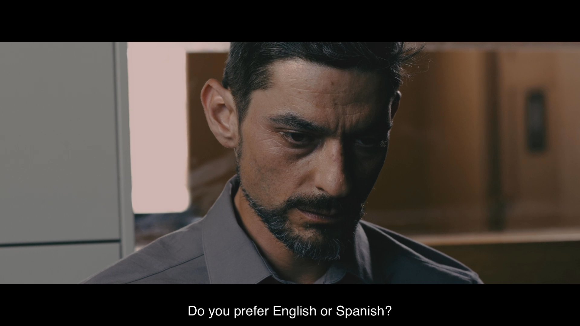 An investigator questions a man named Francisco Carrillo inside a formal setting, beginning with identity verification and language preference. The atmosphere remains tense as the formal interview proceeds.
