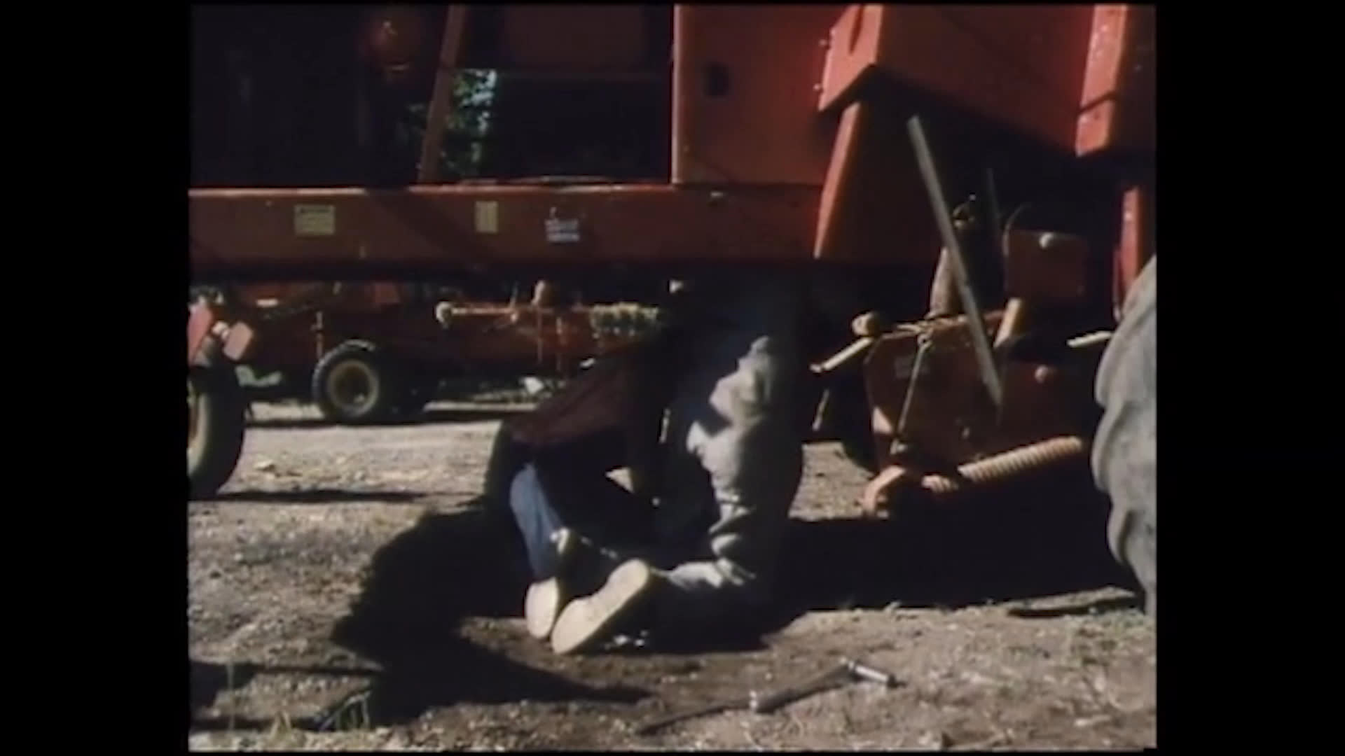 A worker in a cap and overalls crouches beside red farm machinery, then reaches beneath it to investigate something inside. Another person remains low near the equipment as they discuss what may be underneath.
