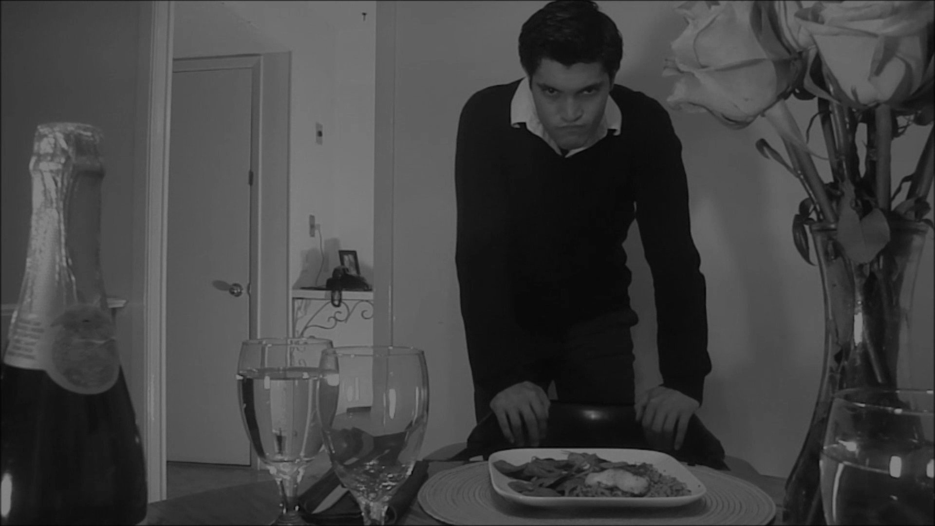 A man enters a dining room, approaches a table set for a meal, and sits down before reaching for his plate. The scene conveys a quiet, domestic moment captured in a monochrome aesthetic.
