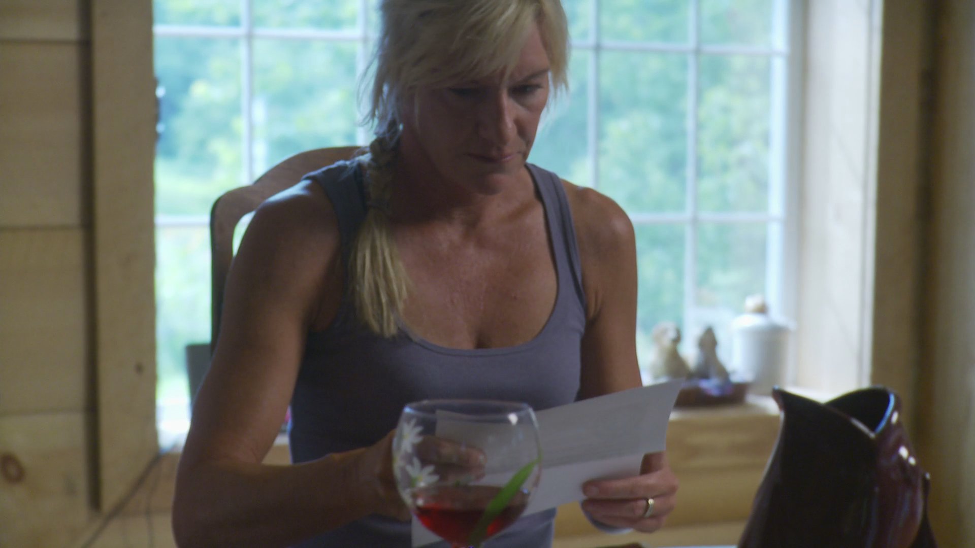 A woman sits at a table in a sunlit room, carefully reviewing a document while holding a glass of wine.