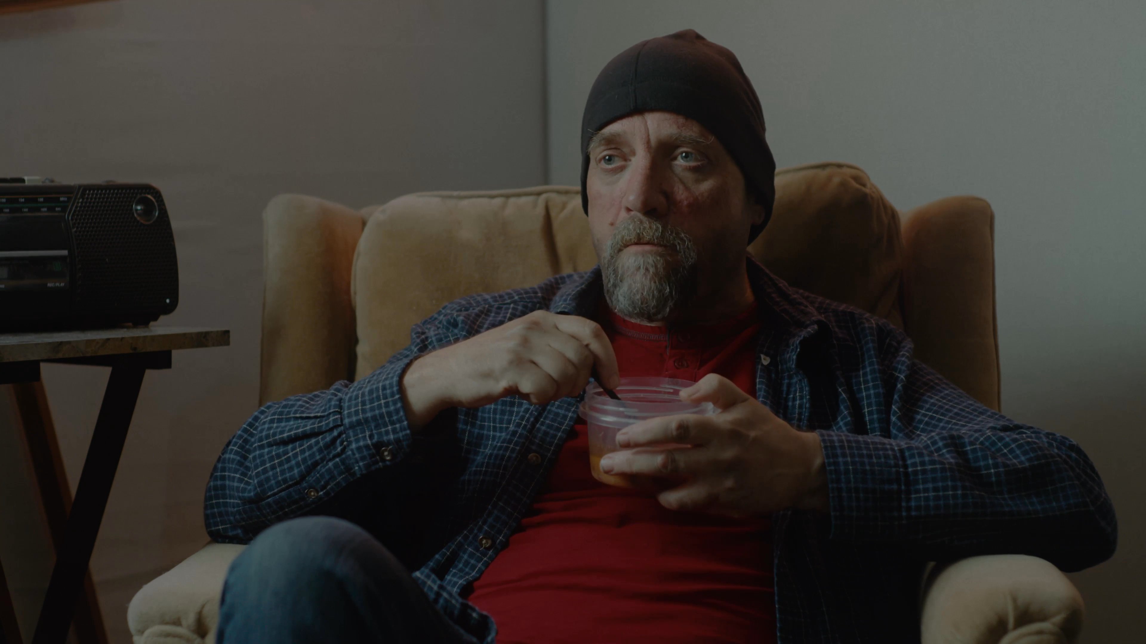 Two men engage in a relaxed conversation while sitting in a living room, with one individual eating a meal from a plastic container.