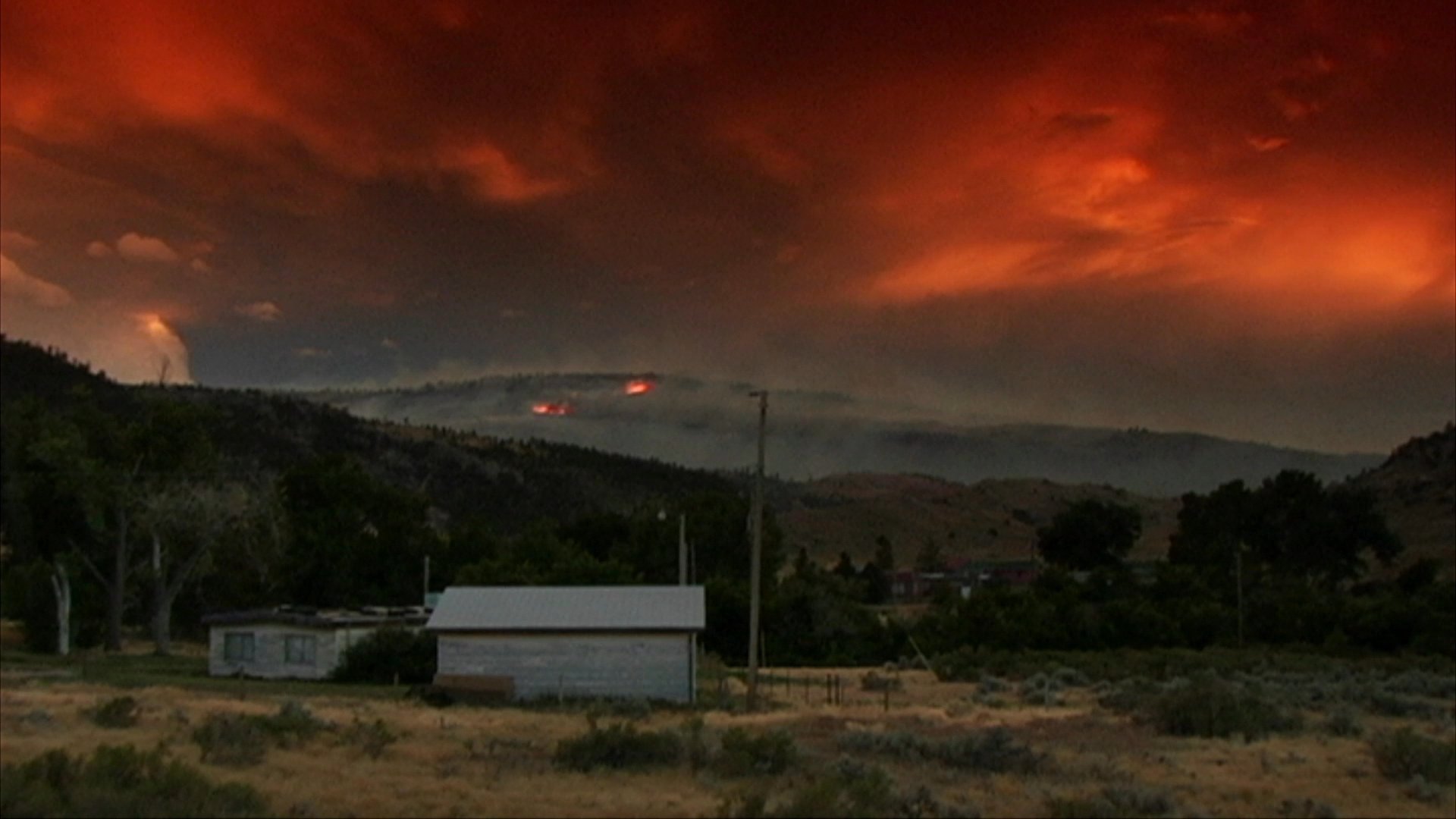 A group of motorcyclists rides along a scenic road at dusk, contrasting with a distant hillside engulfed in wildfire flames. The atmospheric scene transitions from a rainy vehicle interior to the illuminated highway under a burning horizon.