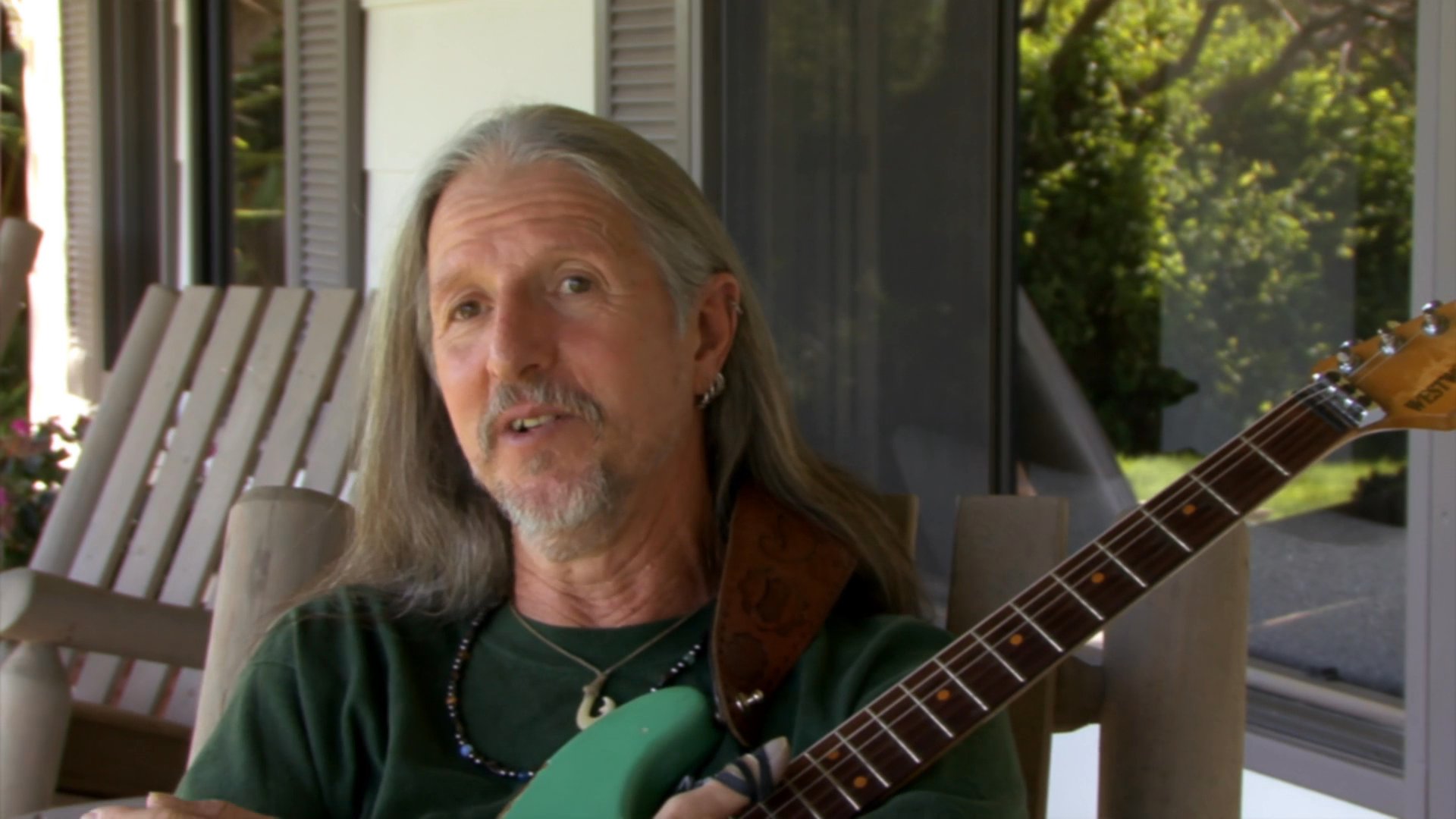 A seasoned musician sits outdoors with an electric guitar, sharing insights through expressive storytelling and hand gestures. He speaks directly to the camera in a relaxed, conversational manner.