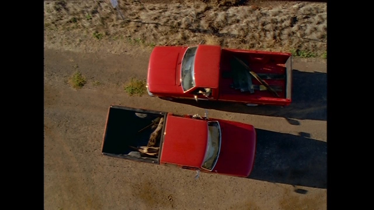 Two men engage in a discussion while traveling in a red pickup truck along a coastal path near a bridge structure. The scene shifts from an overhead perspective of the vehicle to a close-up of the passengers talking inside the cabin.