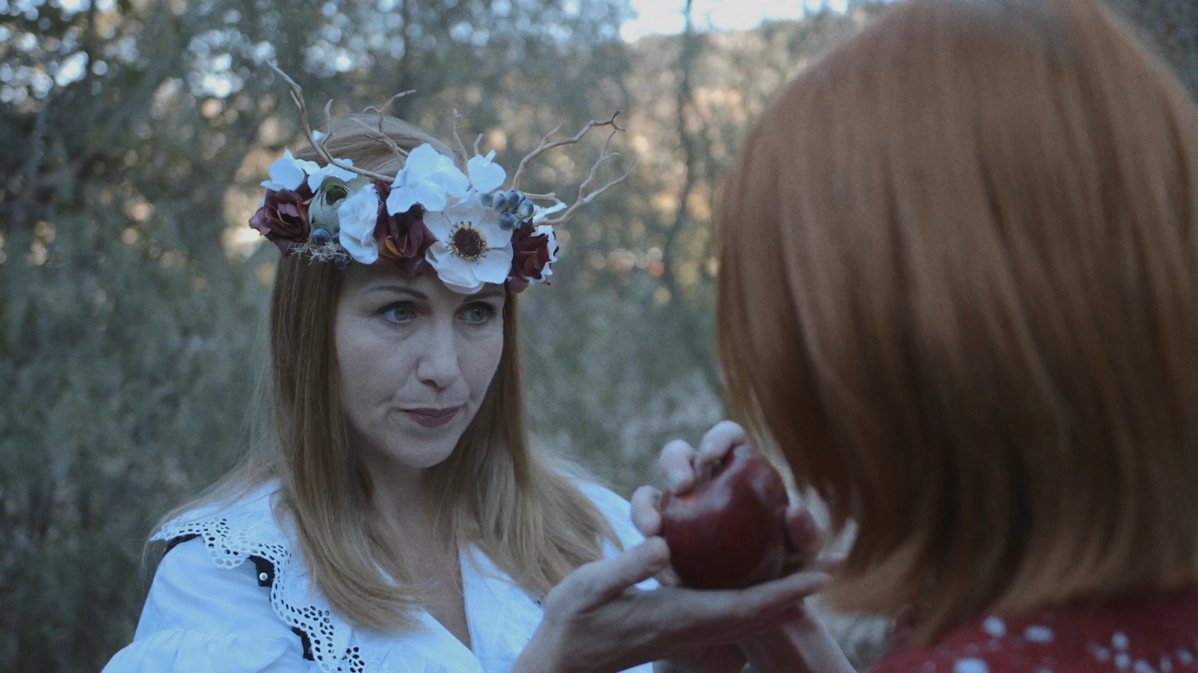 A woman wearing a floral wreath offers a dark red apple to a younger woman in a red dress within a forest setting. The scene evokes a classic fairytale narrative as the recipient hesitates before accepting the fruit.