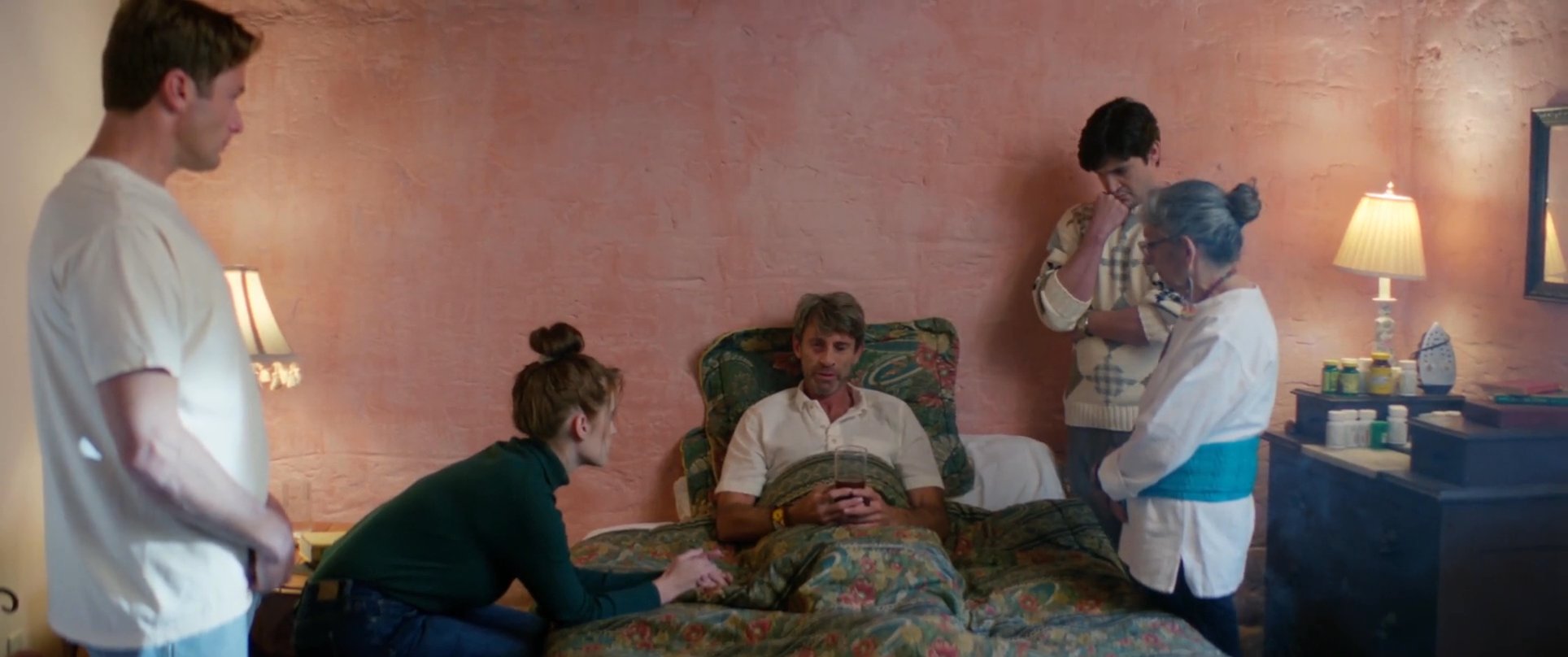 A group of family members gathers around a man resting in bed, offering support and care in a somber, quiet environment. The scene captures a moment of shared concern and emotional connection during a time of health challenges.