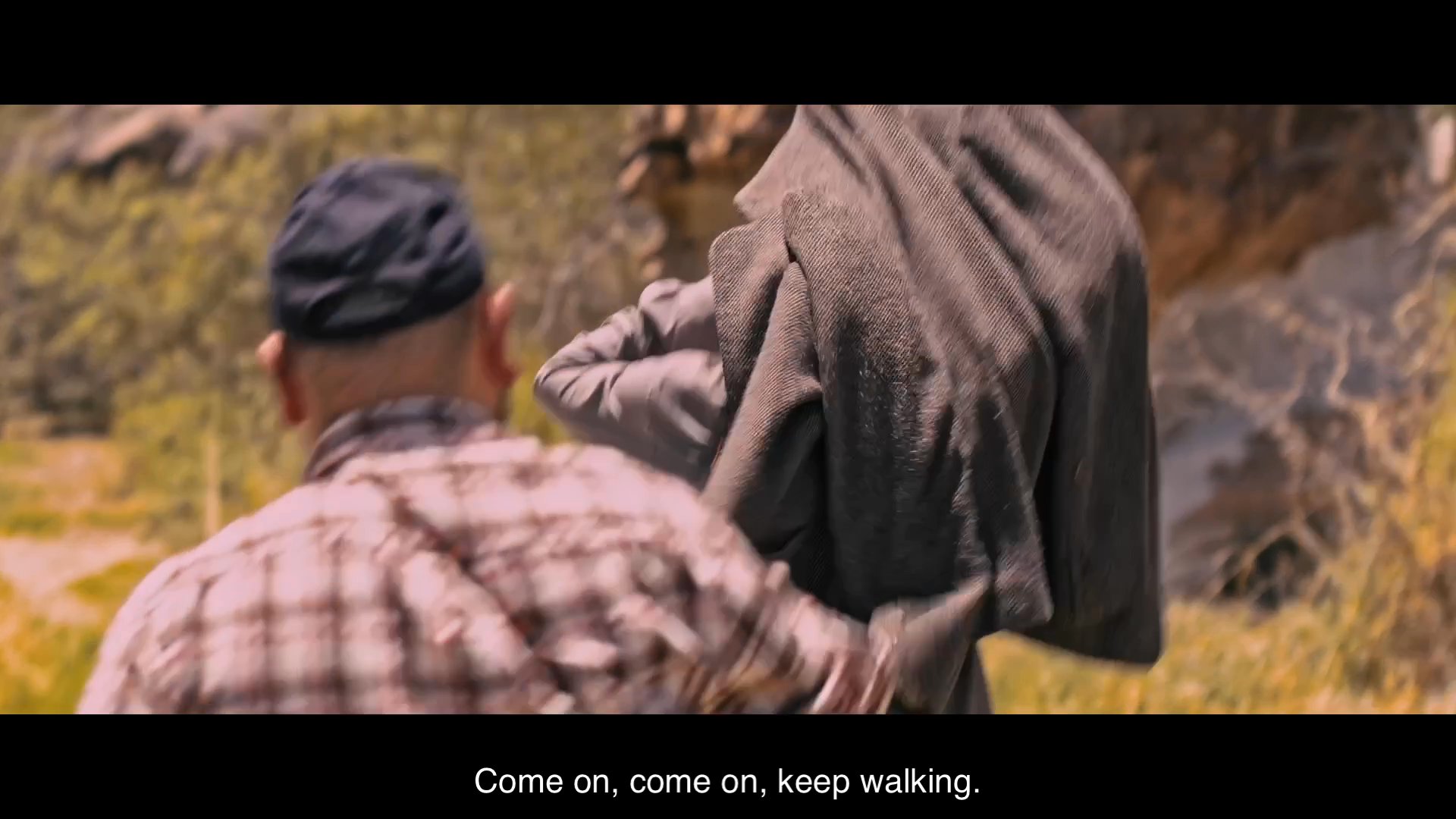 Two men navigate through a harsh, arid desert landscape while discussing their route. One individual leads the way, urging his companion to continue walking as they traverse the barren terrain.