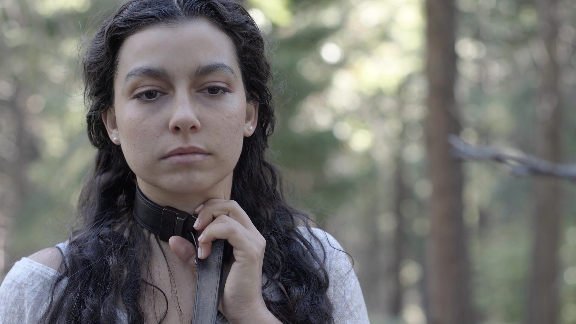 A woman stands in a forest setting and carefully fastens a black leather choker around her neck.