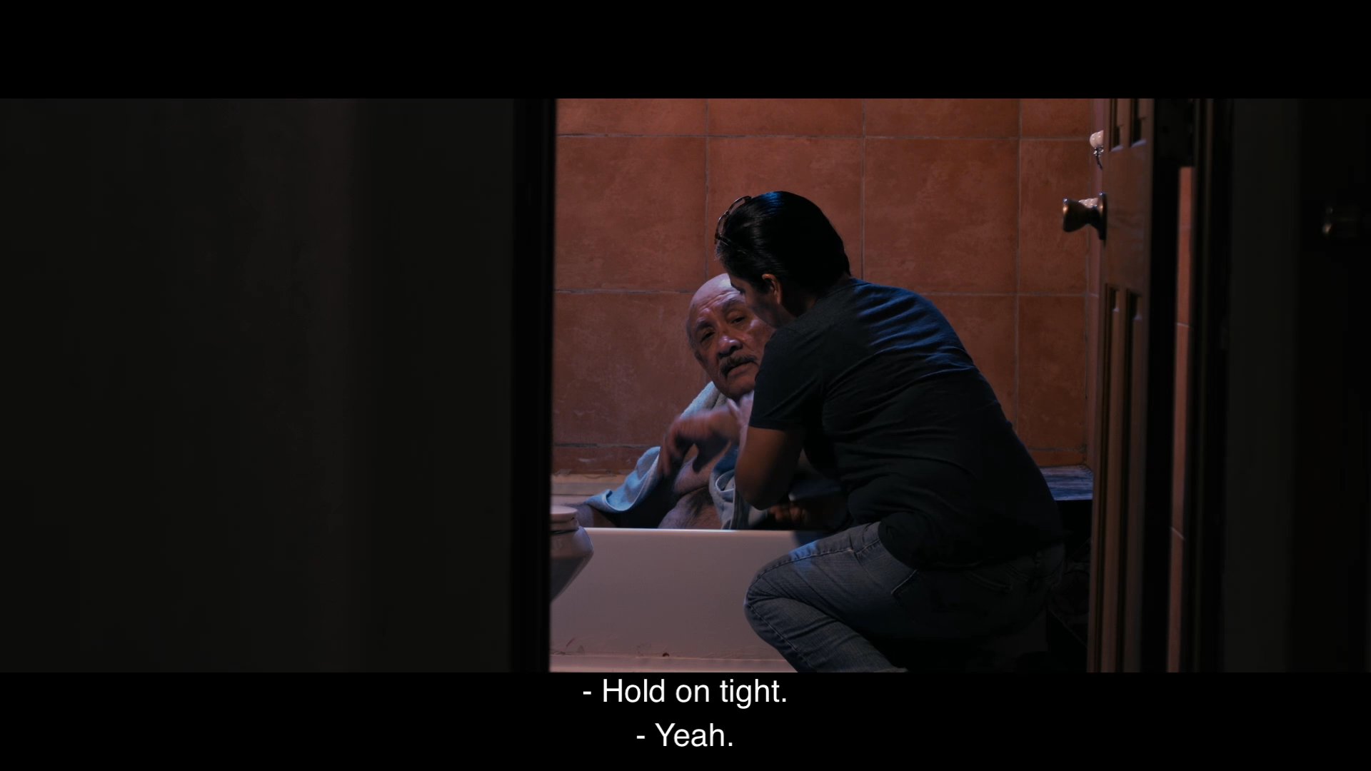 A caregiver assists an elderly man in exiting a bathtub and helps him walk slowly out of the bathroom with support. The scene highlights the patient coordination required during personal care and mobility assistance.