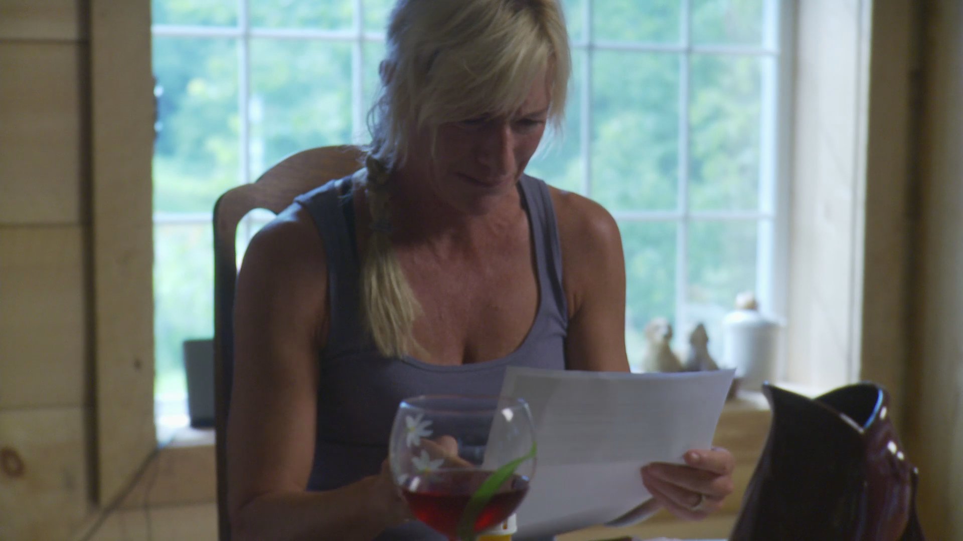 A woman sits alone in a kitchen, visibly upset while reading a document and drinking wine. Her emotional reaction suggests distressing news contained within the letter.