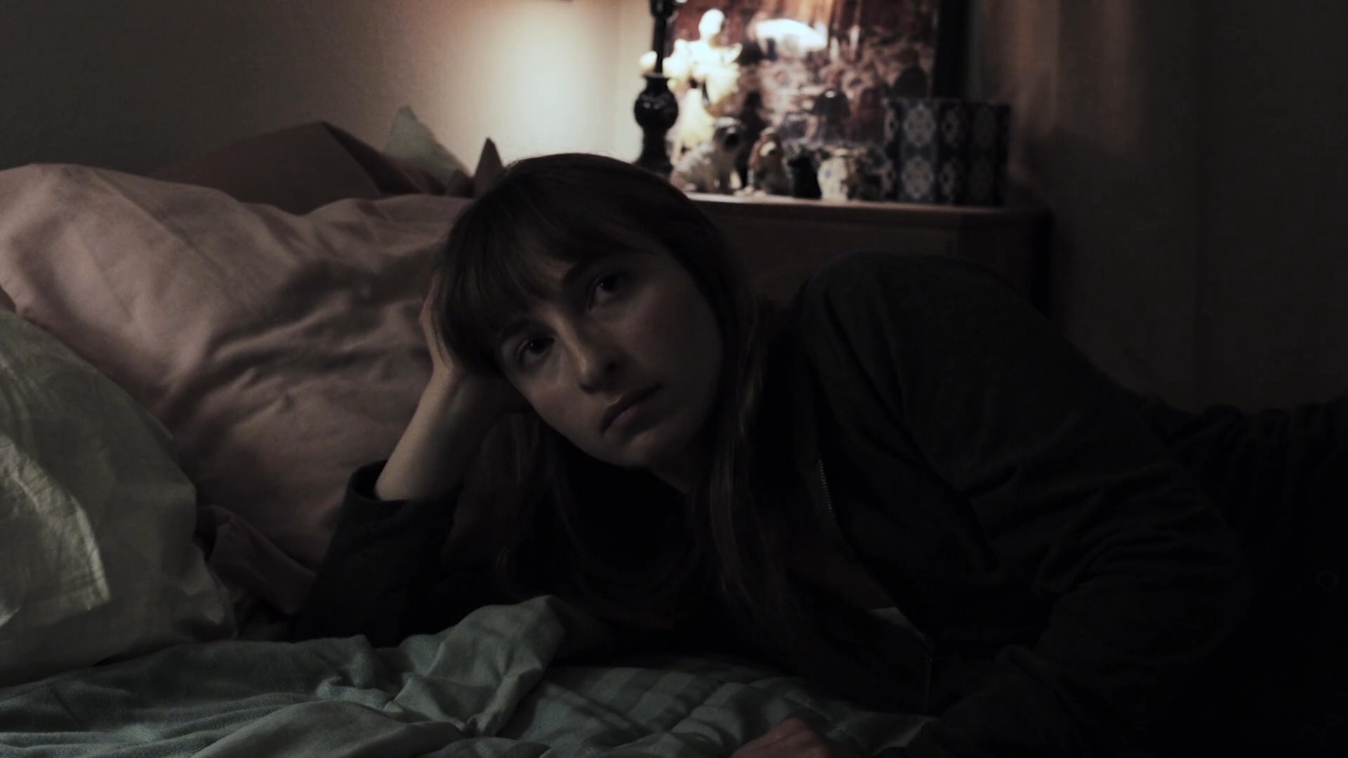A woman enters a dimly lit bedroom, slowly crawls onto the bed, and gazes contemplatively at a landscape print before eventually lying down to rest.
