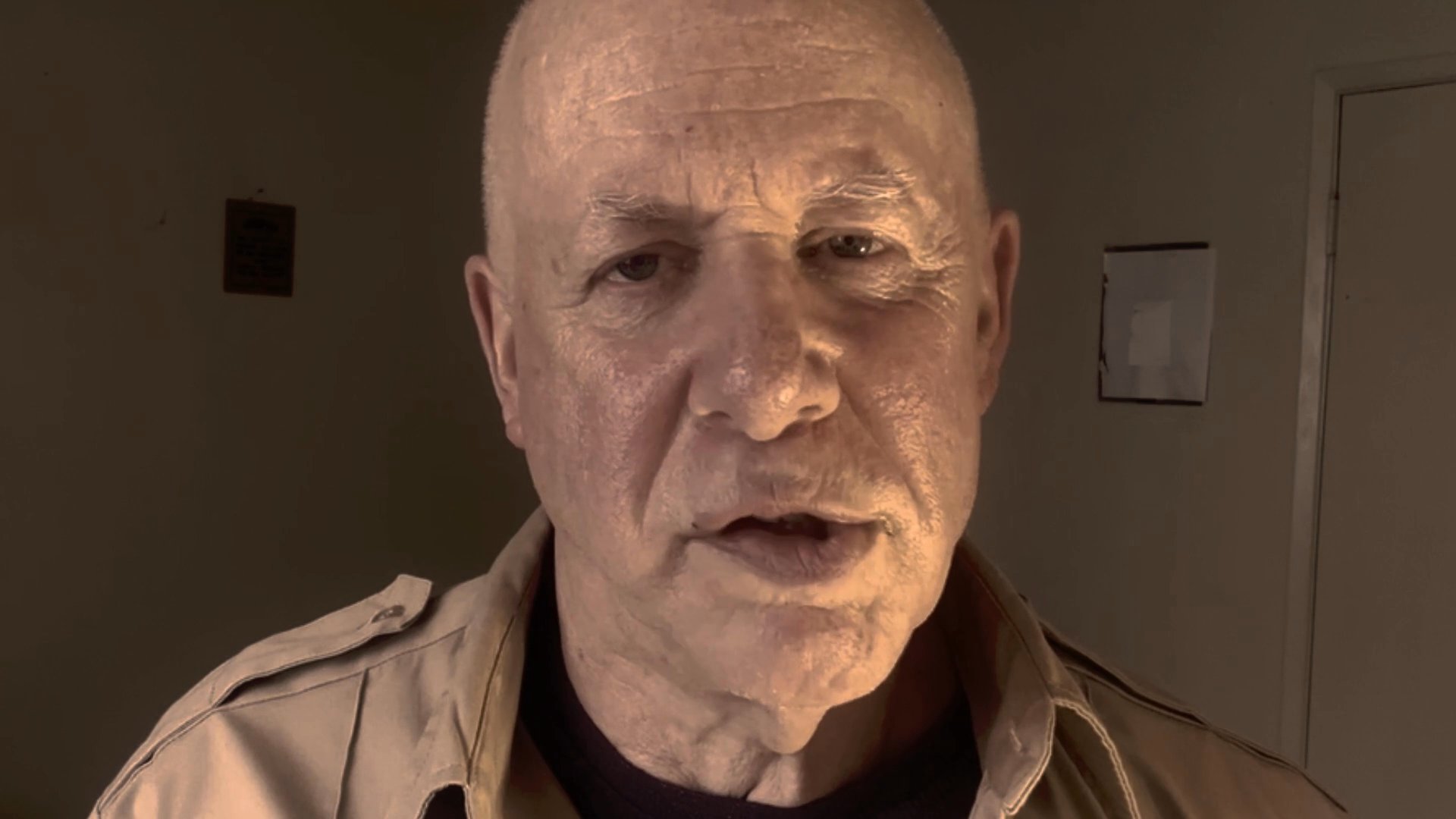 An older man addresses the viewer directly, speaking with a serious and focused expression in a dimly lit environment.