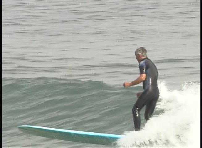 A surfer rides a longboard across ocean waves, followed by an interview segment featuring a man discussing his experience.
