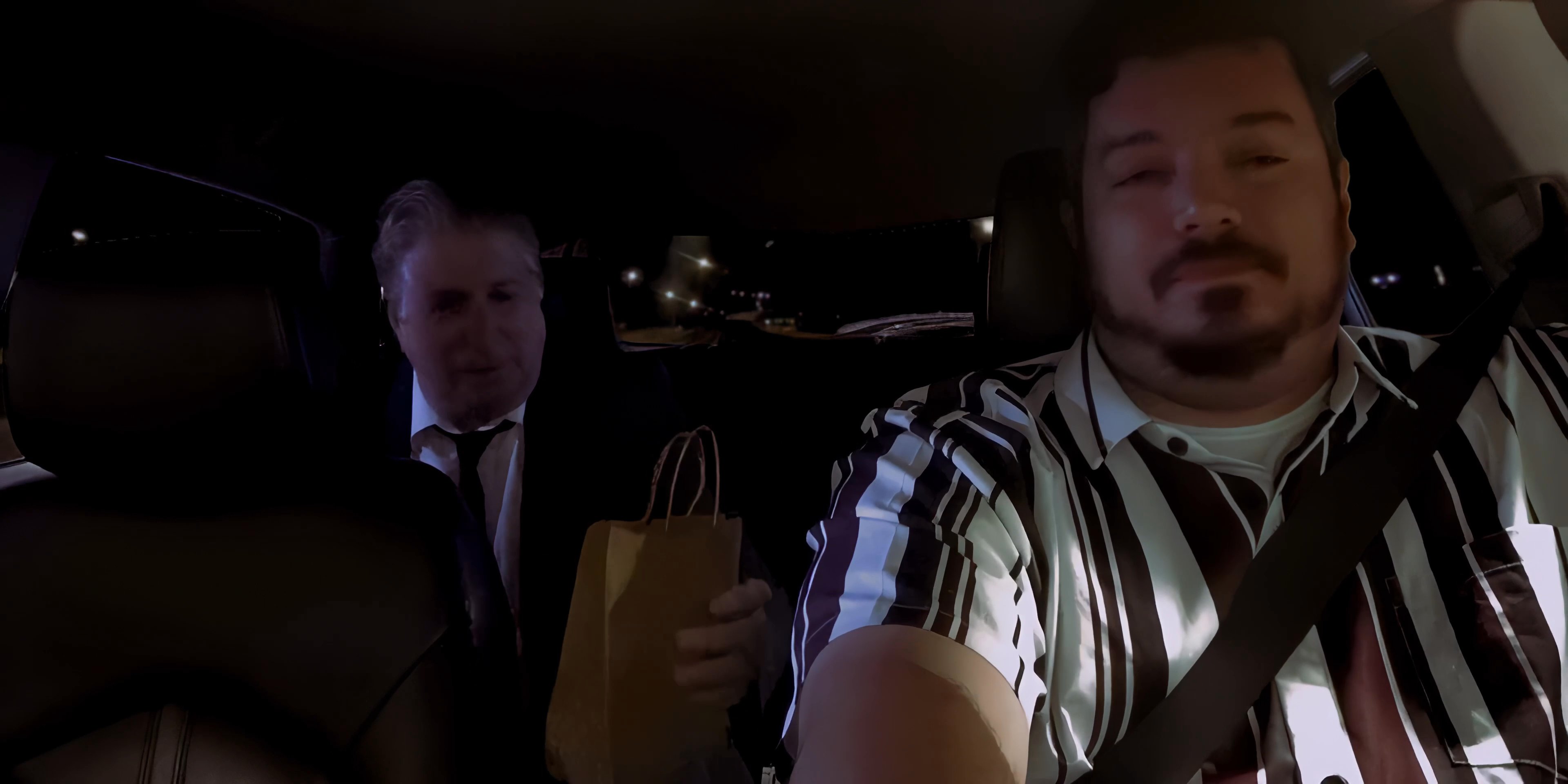 Two men converse in a vehicle at night, with one passenger handing a small gift bag to the driver.