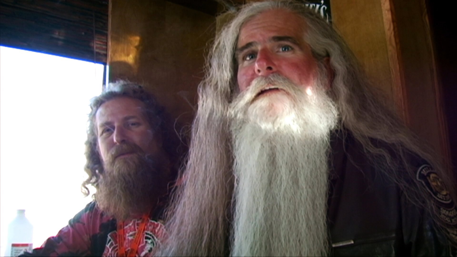 A musician with a long white beard engages in a conversation before transitioning to an acoustic guitar session. The setting appears to be a casual, possibly backstage or studio environment.