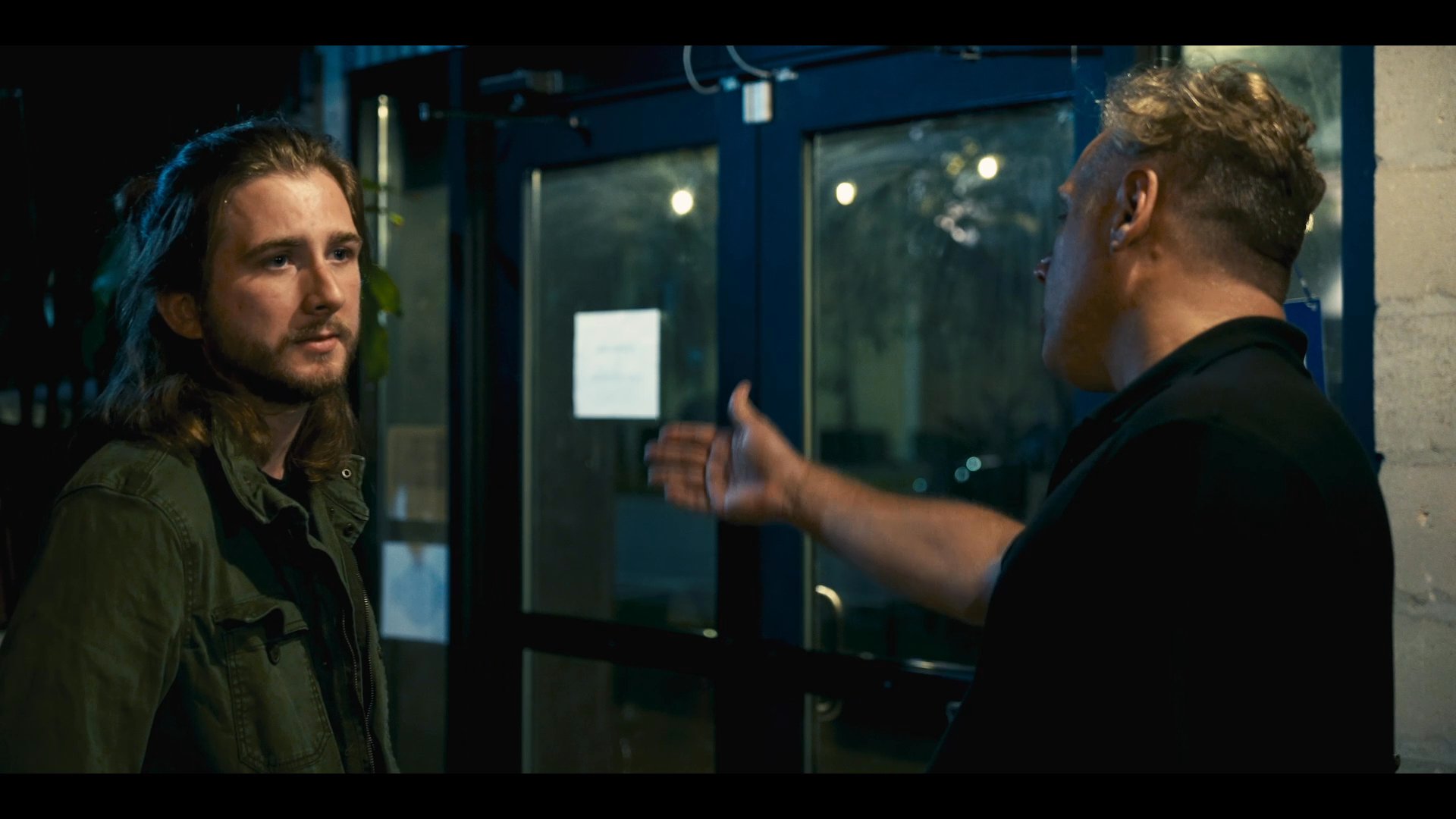 Two couples engage in separate interactions outside a commercial building during the evening. One pair embraces near a wall while another pair holds a focused conversation near the entrance.
